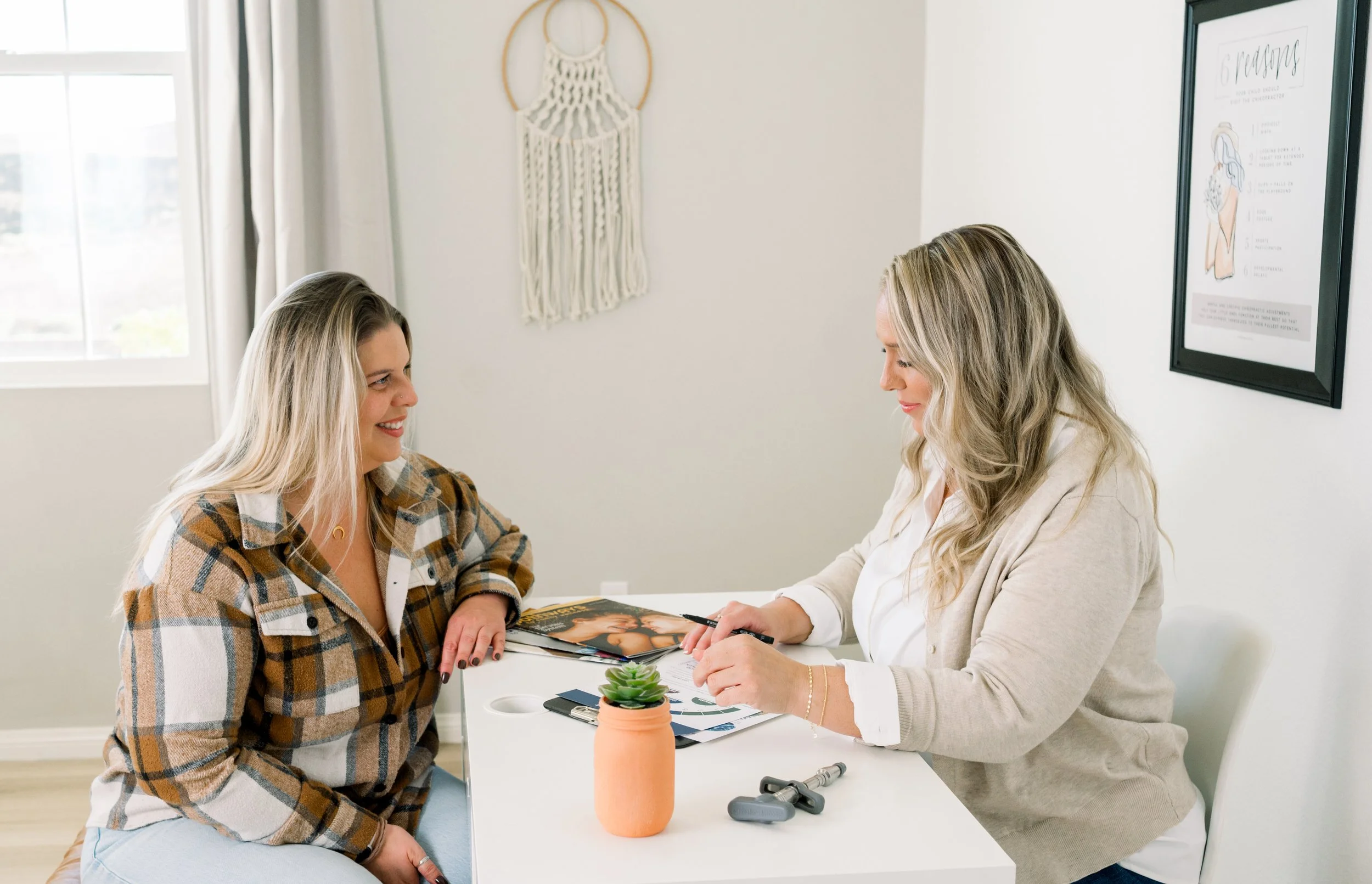 A woman sitting across from another woman at a table during a consultation, with various papers, a small plant in a terracotta pot, and a chiropractic tool on the table. The setting appears to be a bright, modern consultation room.