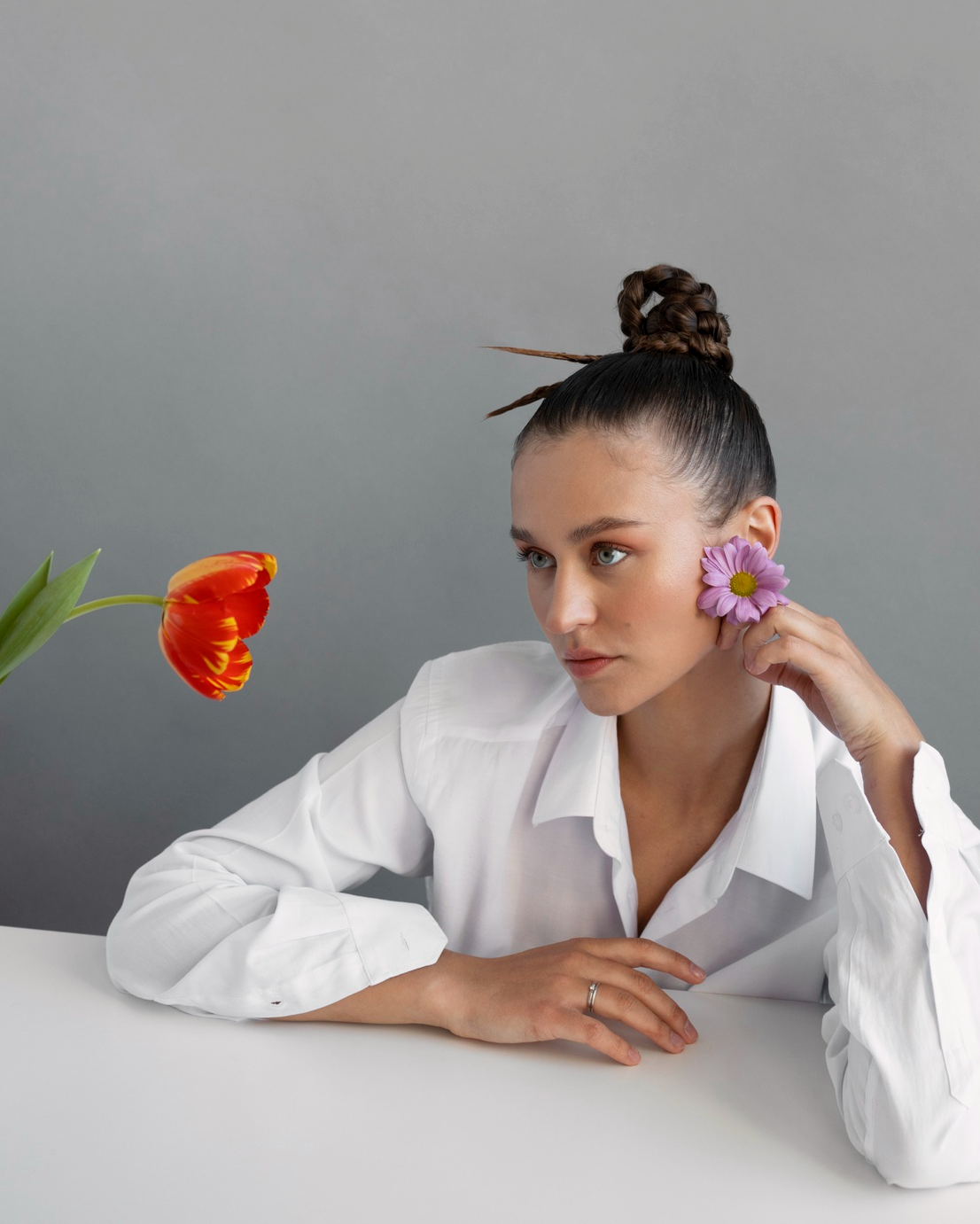 A woman with dark hair styled in a bun with braids, wearing a white shirt, holds a pink flower to her cheek while looking at a tulip on her left against a plain gray background.