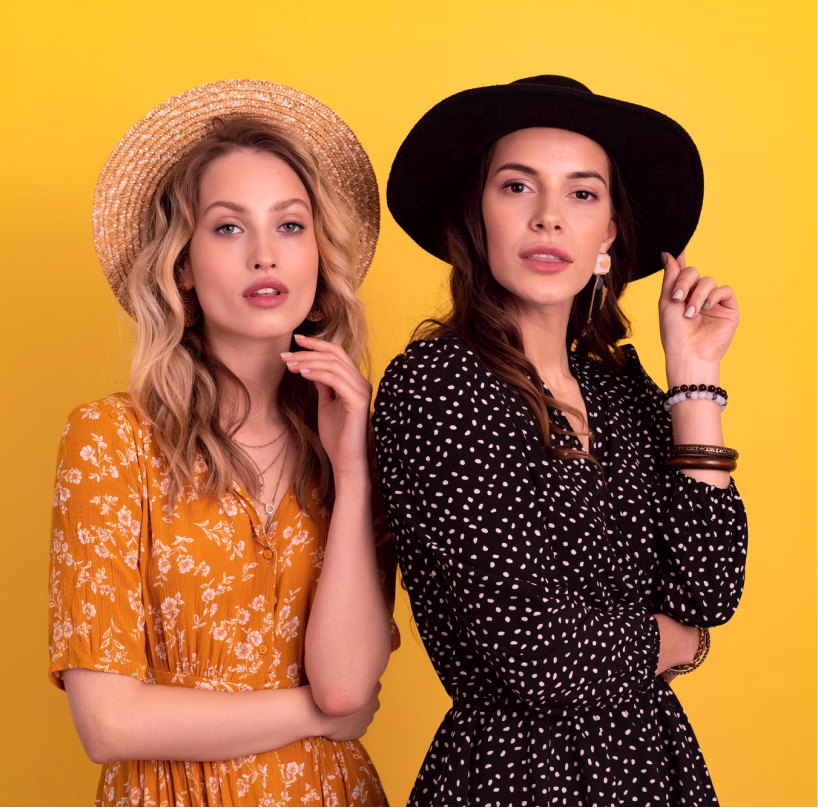 Two women wearing hats and polka dot dresses standing against a yellow background.