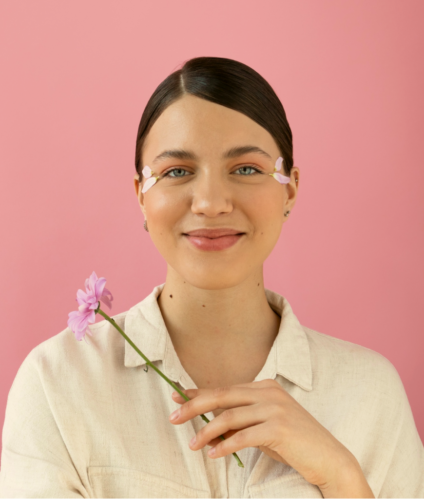 A woman with dark brown hair and blue eyes holding a pink flower against a pink background.