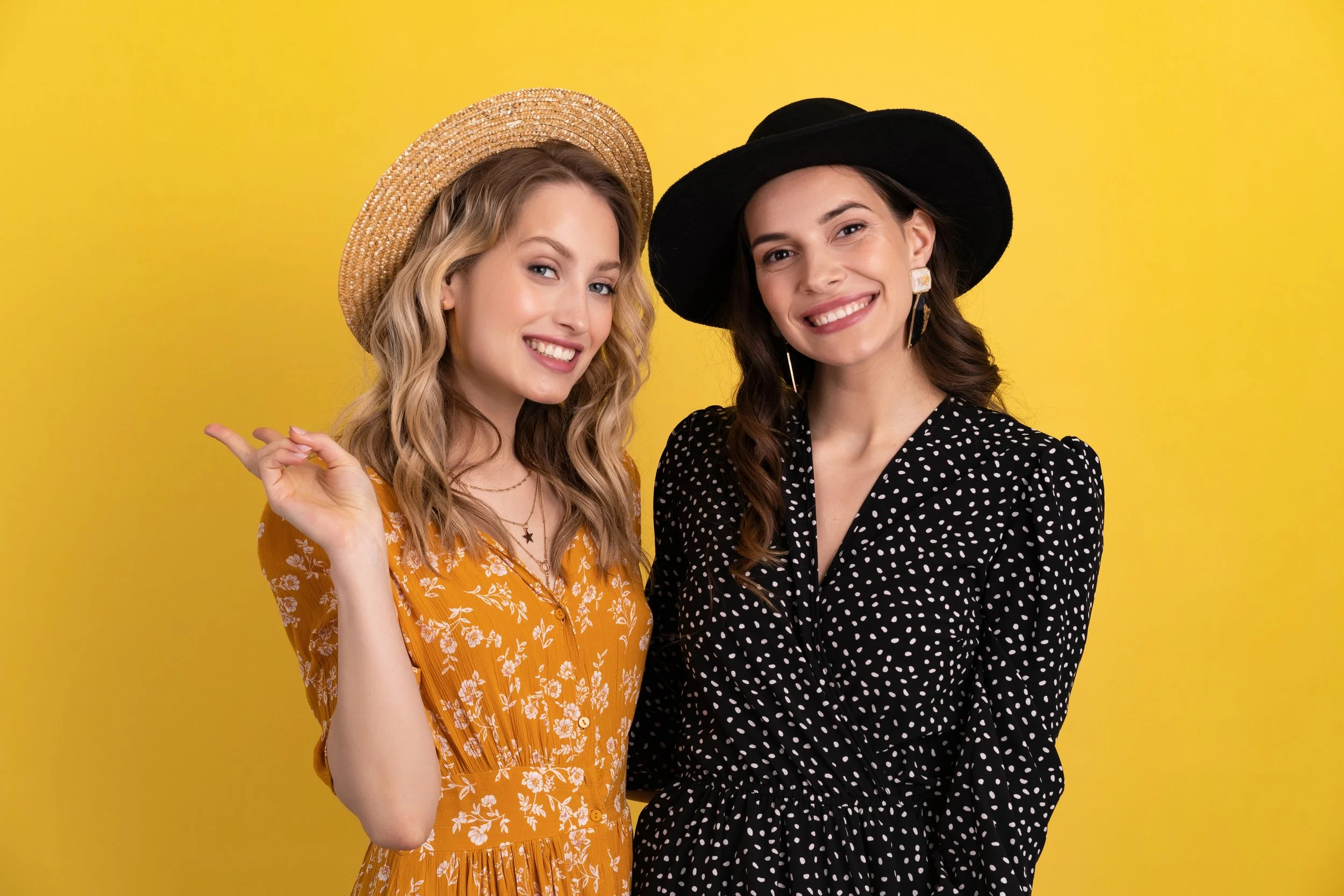 Two women smiling, wearing black and mustard yellow polka dot and floral dresses with wide-brim hats, standing against a yellow background.