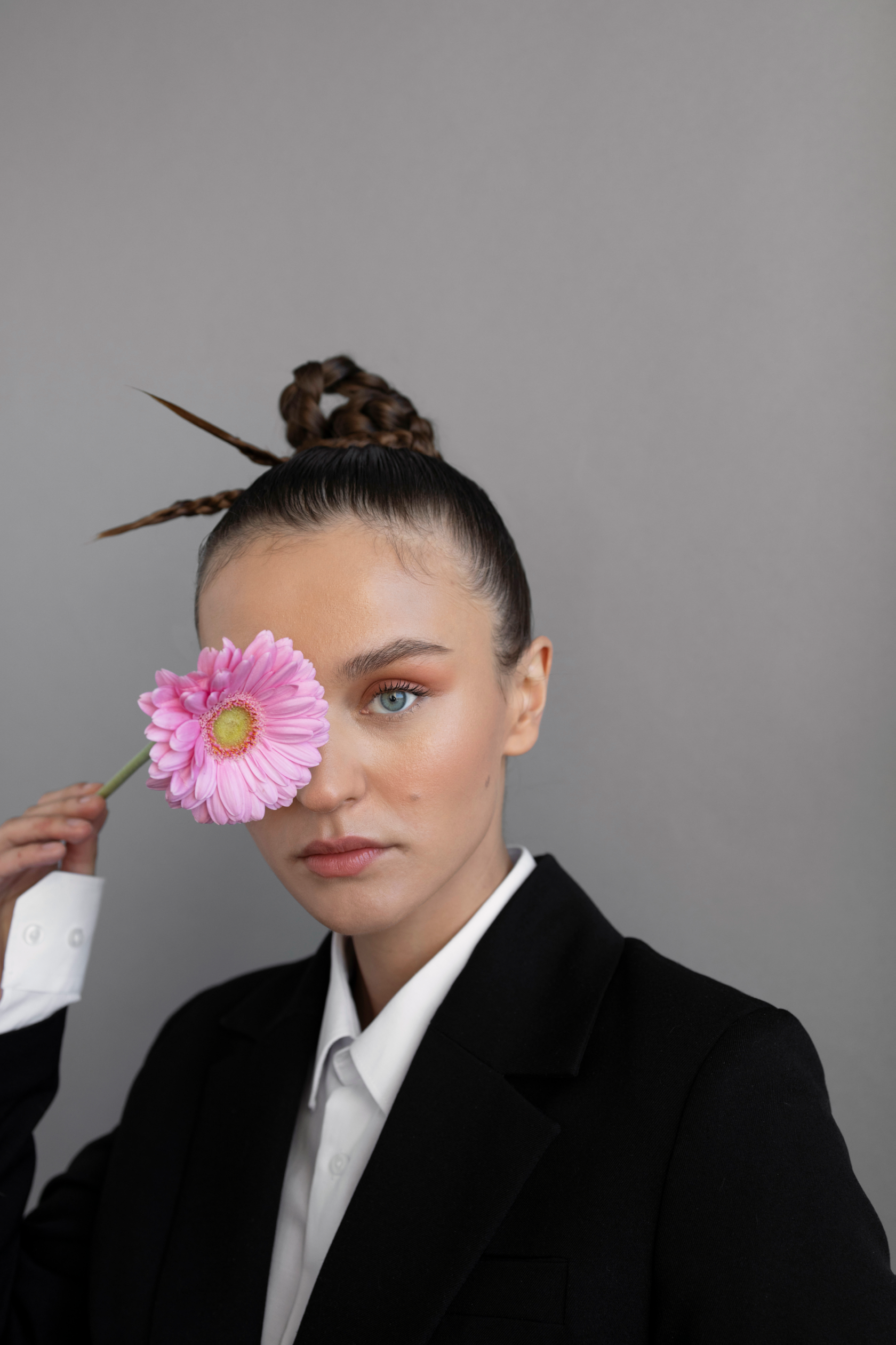 Portrait of a woman holding a pink flower over one eye, with gray background, wearing a black blazer and white shirt.