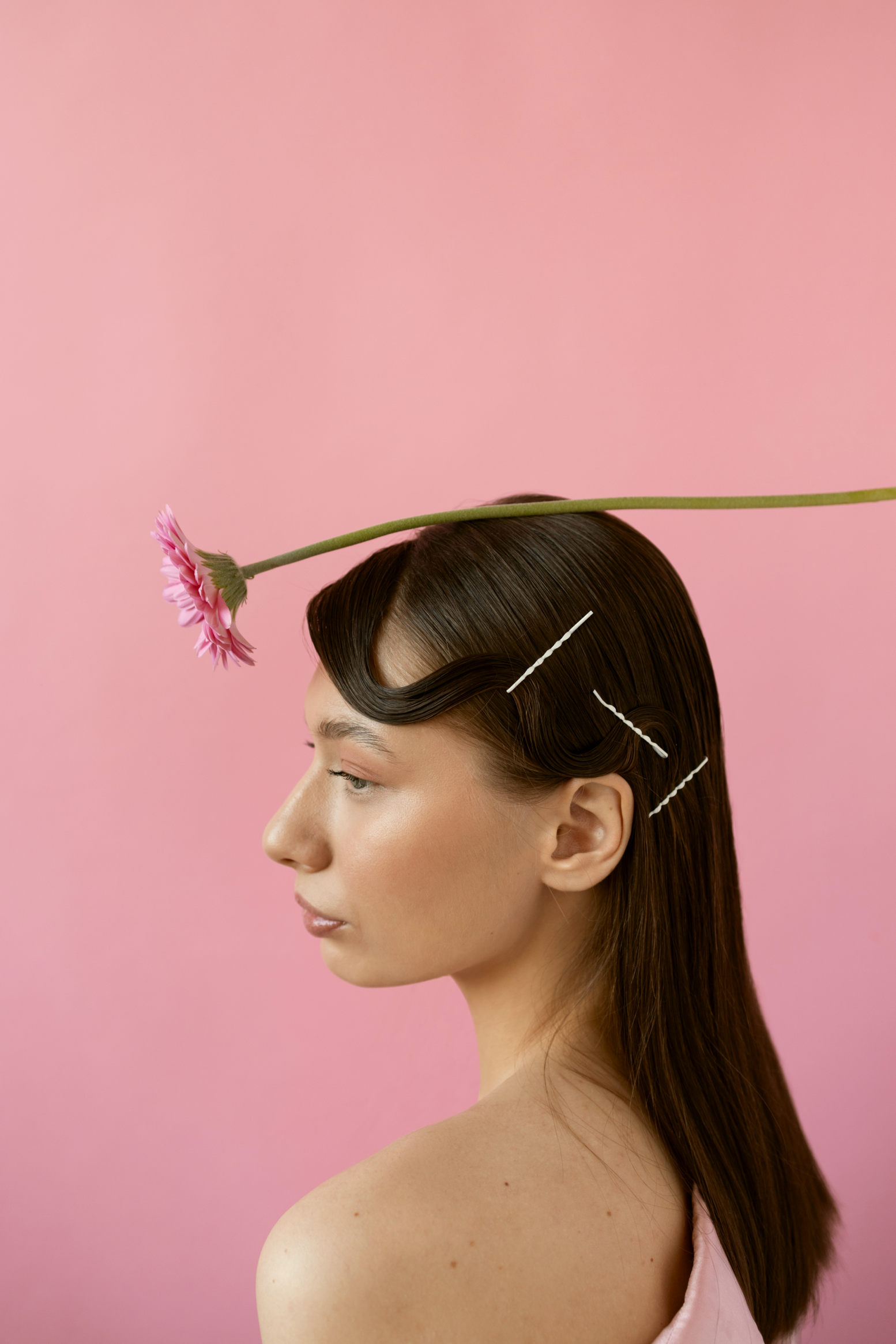 A woman with straight brown hair and styled waves in the front has a pink flower resting on her head, held in place with white hairpins, against a pink background.