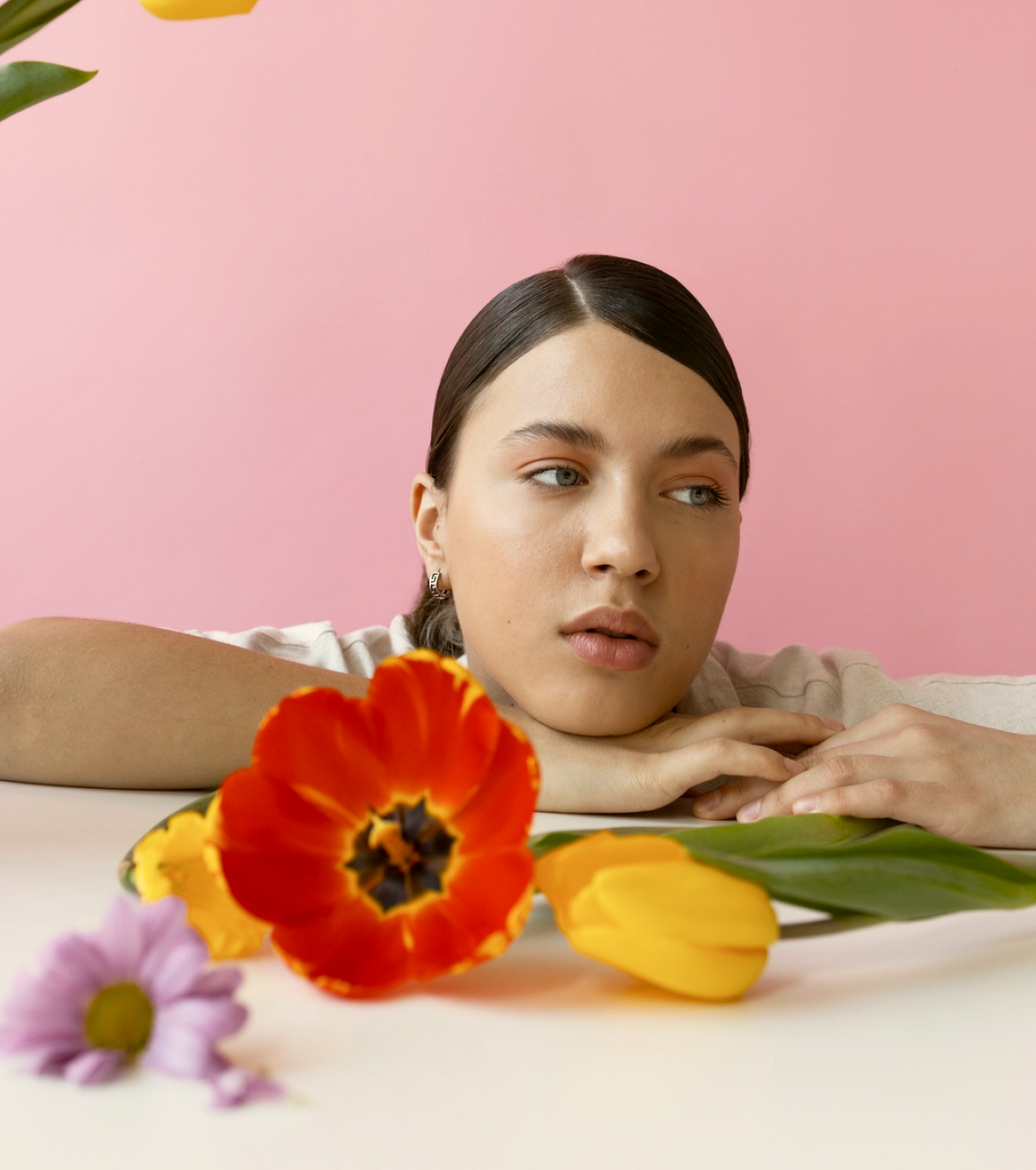 A young woman with dark hair and earrings resting her head on a table, looking to the side with flowers in front of her against a pink background.