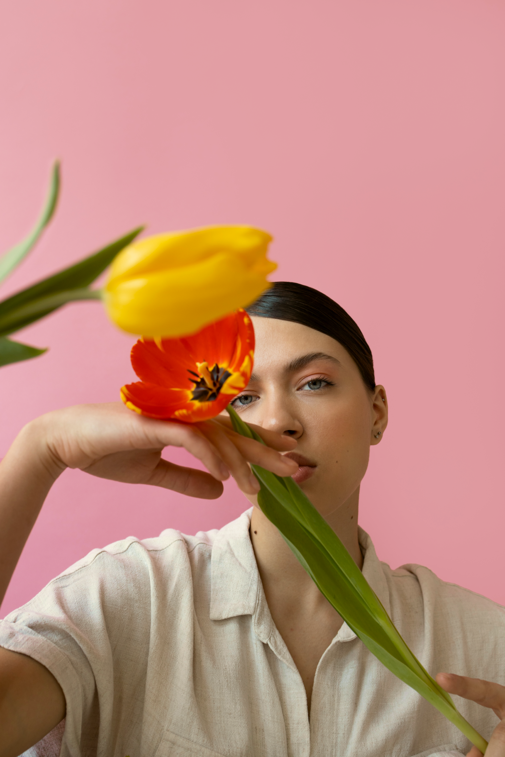 Young woman with dark hair and light skin holding a yellow and red tulip in front of her face against a pink background