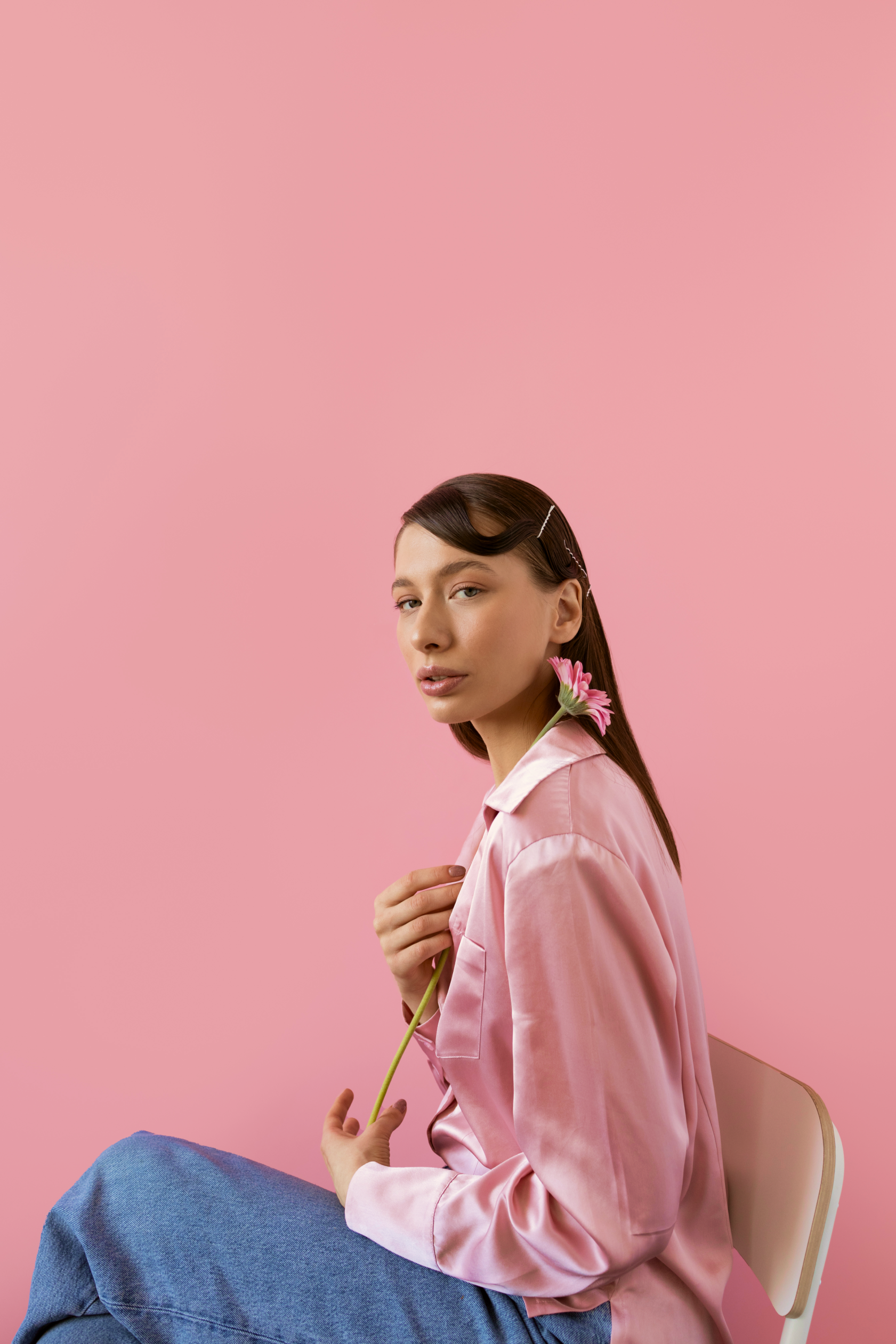 A young woman with brown hair styled with a hair clip, holding a pink flower near her shoulder, wearing a pink silk shirt and blue jeans, sitting against a pink background.