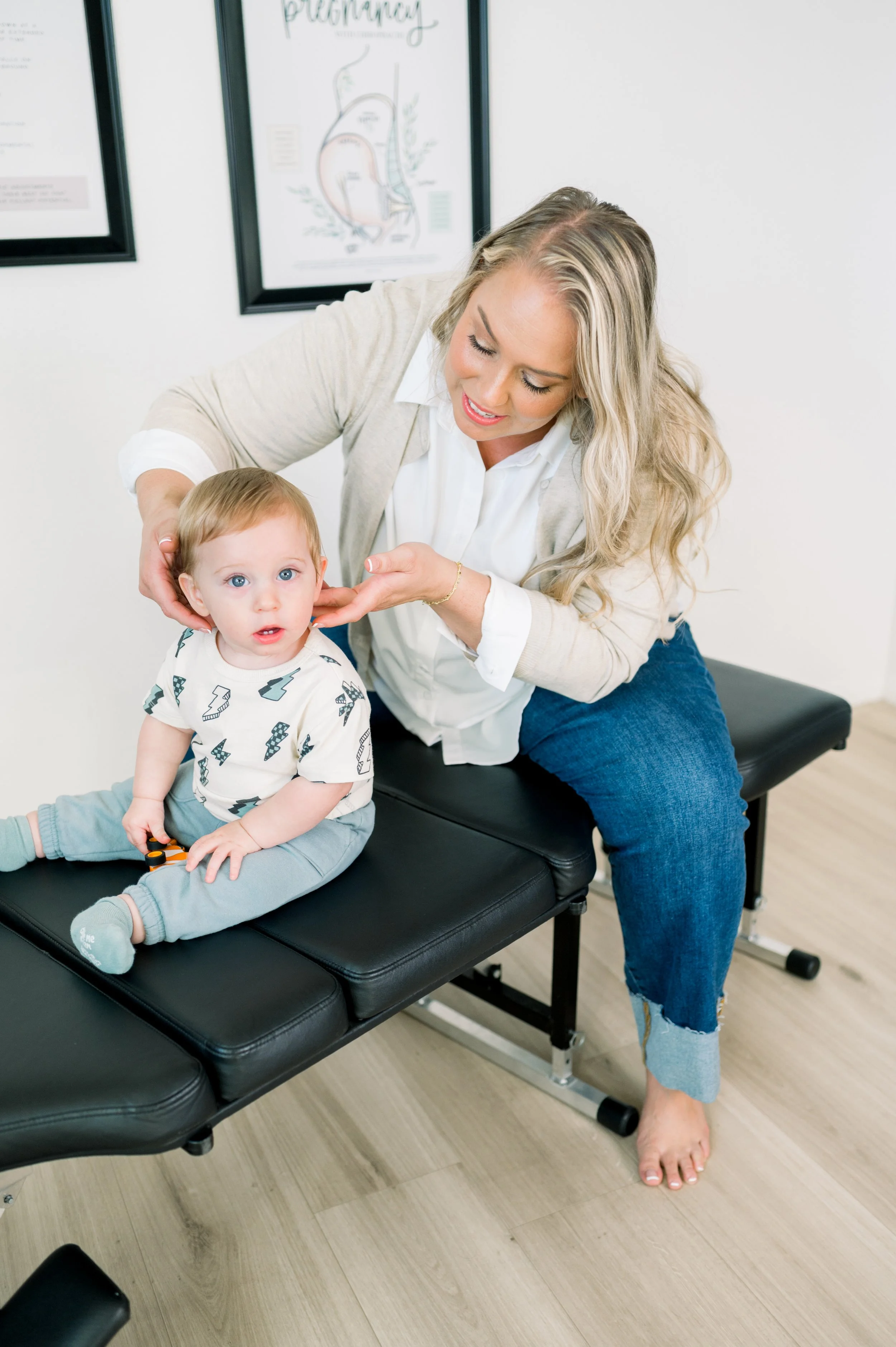 A woman is examining a young child's ear while sitting on an examination table in a medical office. The child is sitting on the table, holding a toy, and looking at the camera. The woman is smiling and gently holding the child's head, with medical posters in the background.