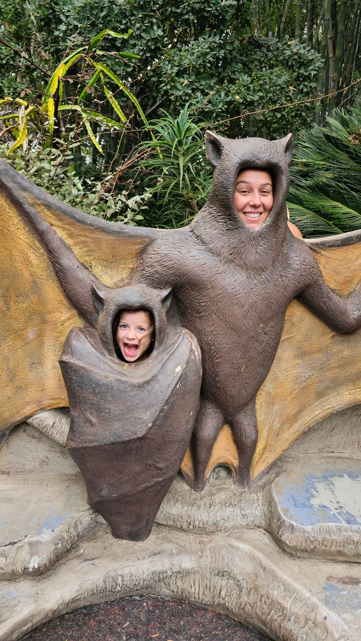 Two children with faces in animal cutout statues of a bat and an owl, in a zoo or park setting.