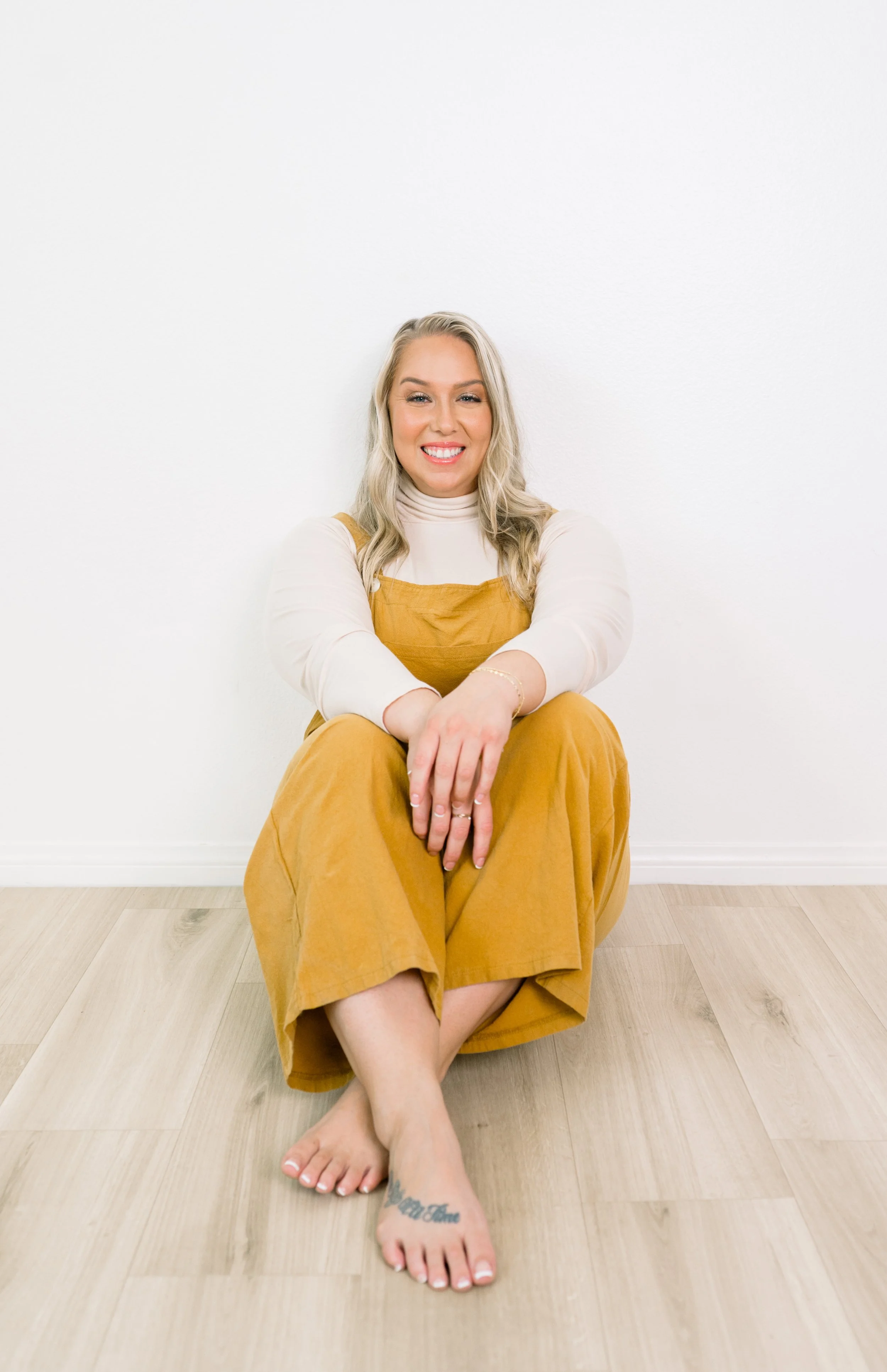 A smiling woman with blonde hair sits on the floor in front of a white wall, wearing a white turtleneck and mustard overalls, with her legs crossed and hands resting on her knees. She has a tattoo on her foot and is barefoot.