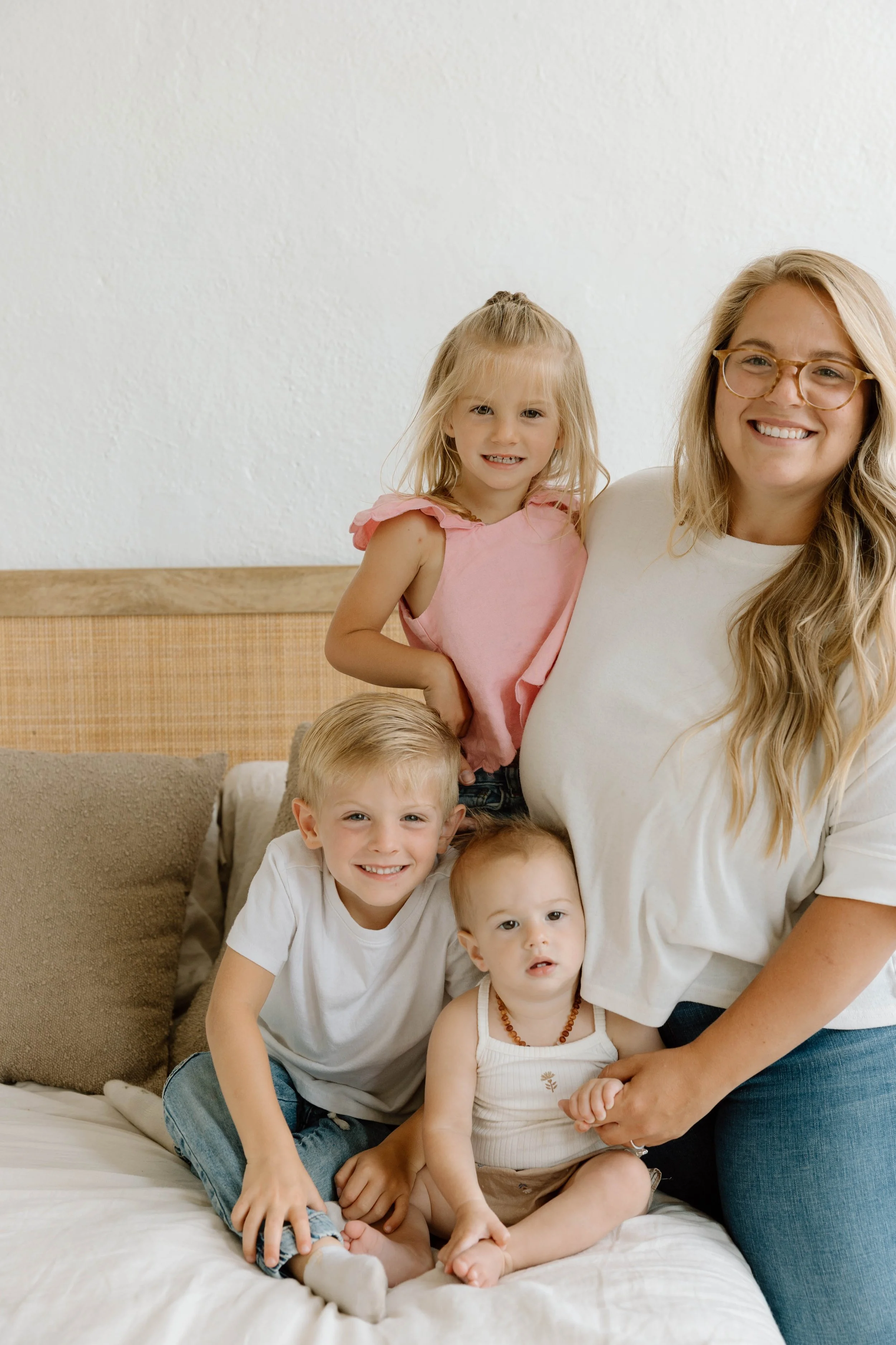 A woman with long blonde hair, glasses, and a white shirt, smiling, with three children on a bed, one girl standing behind her and two boys sitting in front, posing for a photo in a home setting.