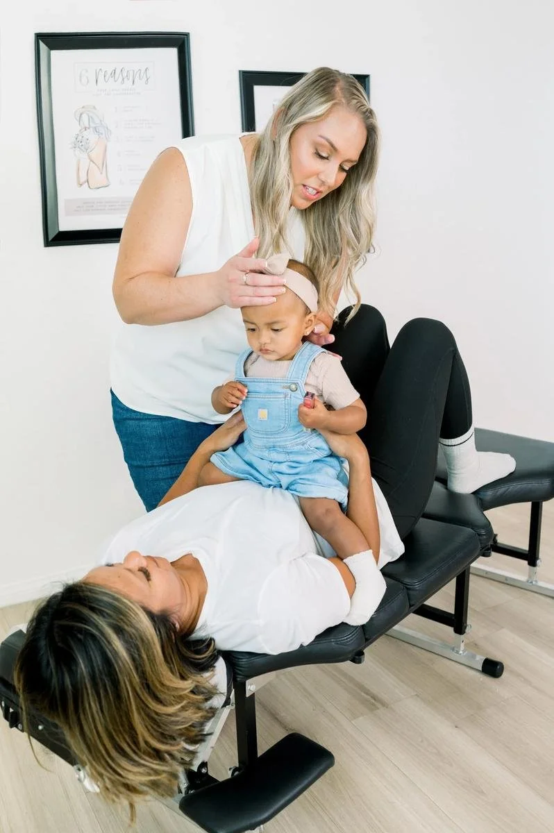 Dr. Jennifer Gordon gently supporting a toddler seated on a parent lying on a chiropractic table.