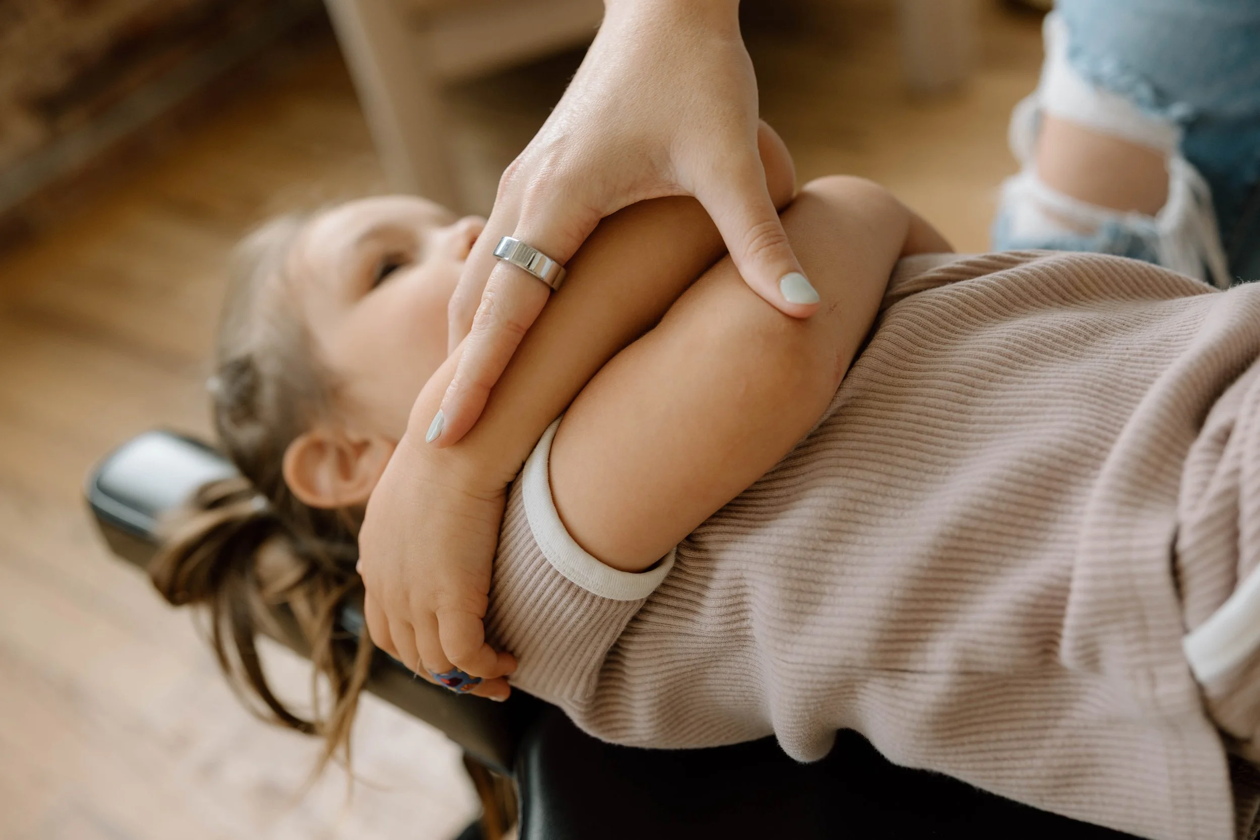 A young girl lying on her back with eyes closed, receiving a shoulder massage from an adult. The girl has light brown hair tied in a ponytail, and is wearing a beige long-sleeve shirt. The adult's hand, wearing a ring, is gently massaging the girl's shoulder. The scene is indoors with wooden flooring visible in the background.