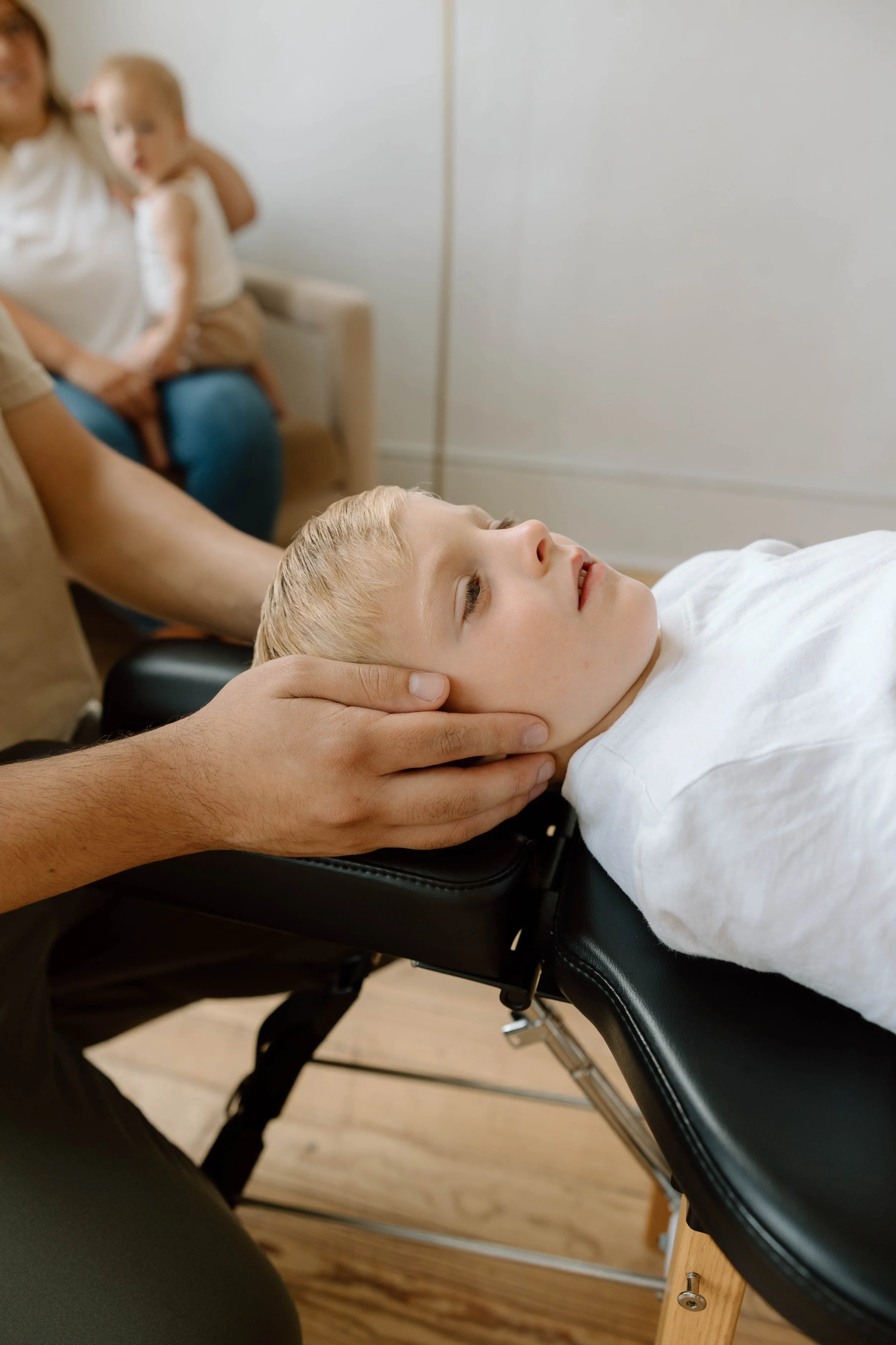 A young boy receiving a chiropractic adjustment while lying face up on a black treatment table, with a practitioner holding his head gently. In the background, a woman holding a toddler is sitting on a beige couch.