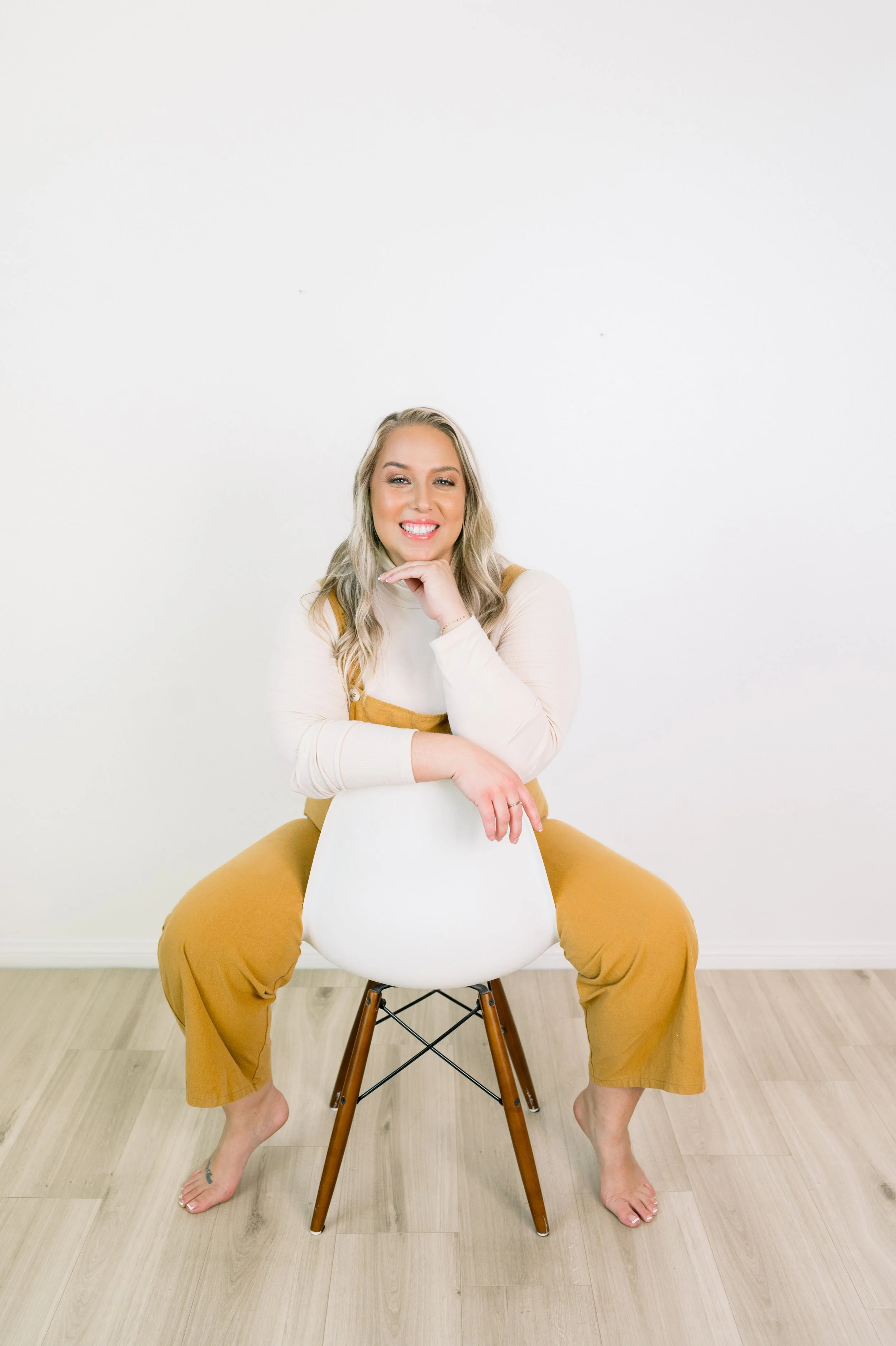 A woman with blonde hair smiling, sitting backwards on a white chair, wearing a cream-colored long sleeve top and mustard yellow overalls, on a wooden floor against a plain white wall.