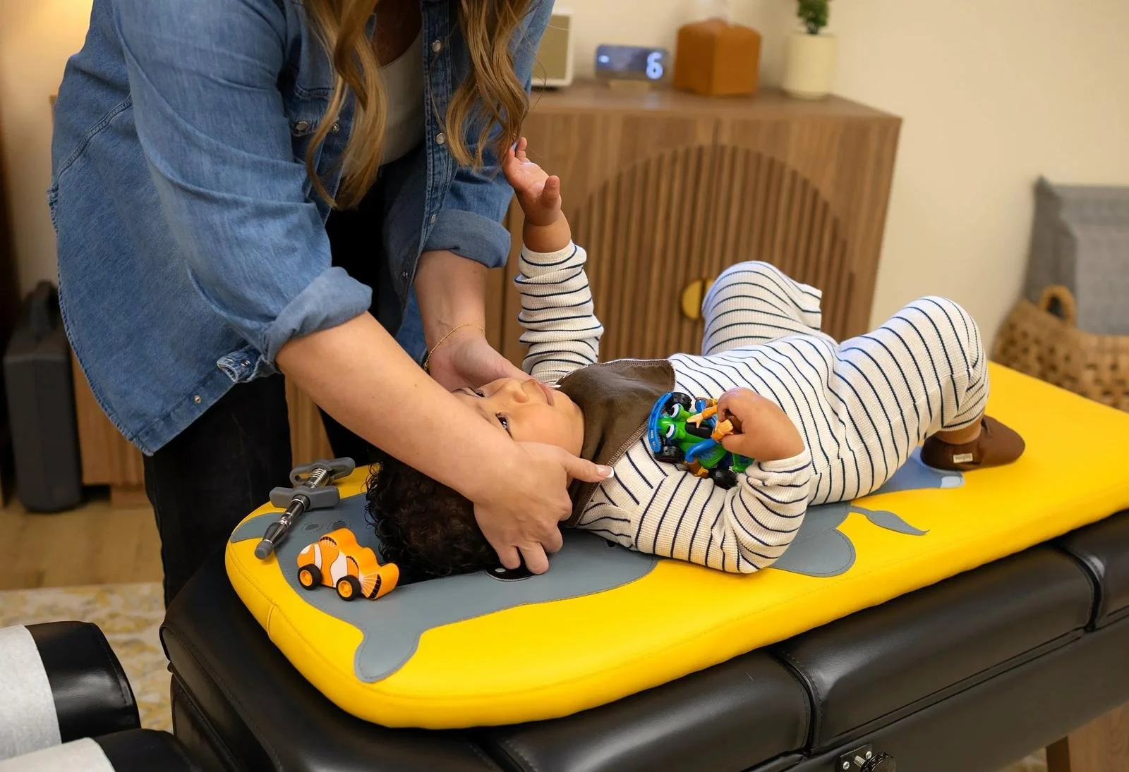 Chiropractor gently supporting a toddler’s neck while the child lies on a pediatric treatment table holding toys.