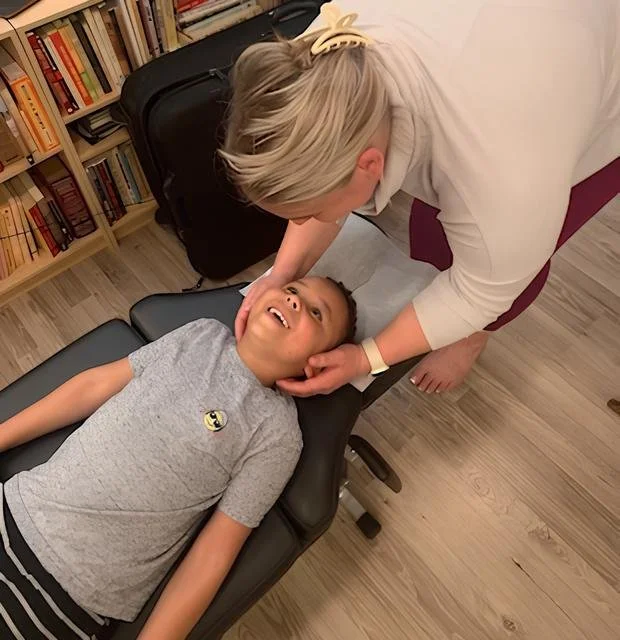 A young boy lying on a black chiropractic table smiling, while a chiropractor, checks his neck in a room with bookshelves and a suitcase in the background.