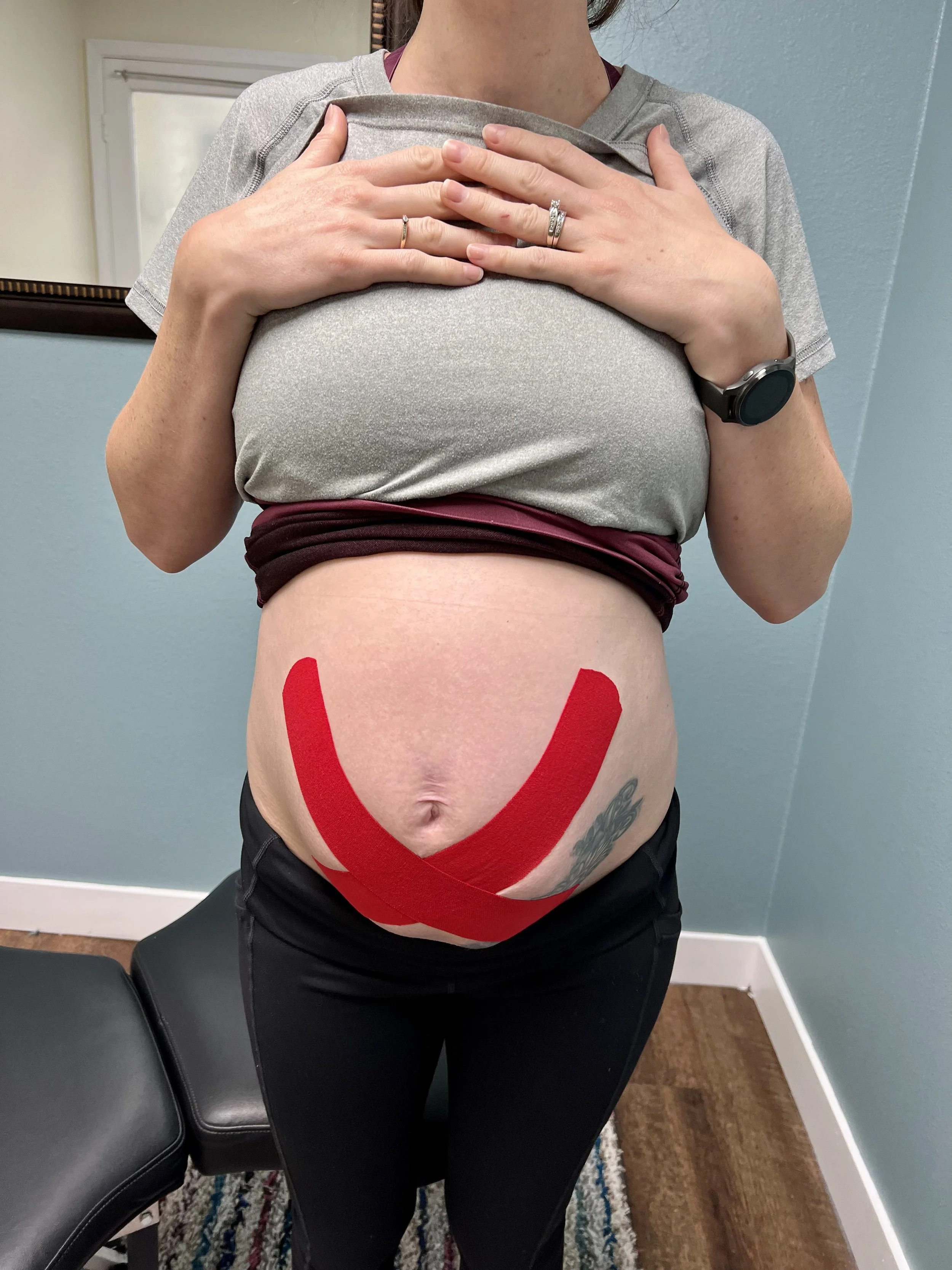 Pregnant woman standing in a chiropractic office, with her grey shirt lifted to show the Kinesiotape taped across her pregnant belly in an 'X' fashion to help support her ligaments.