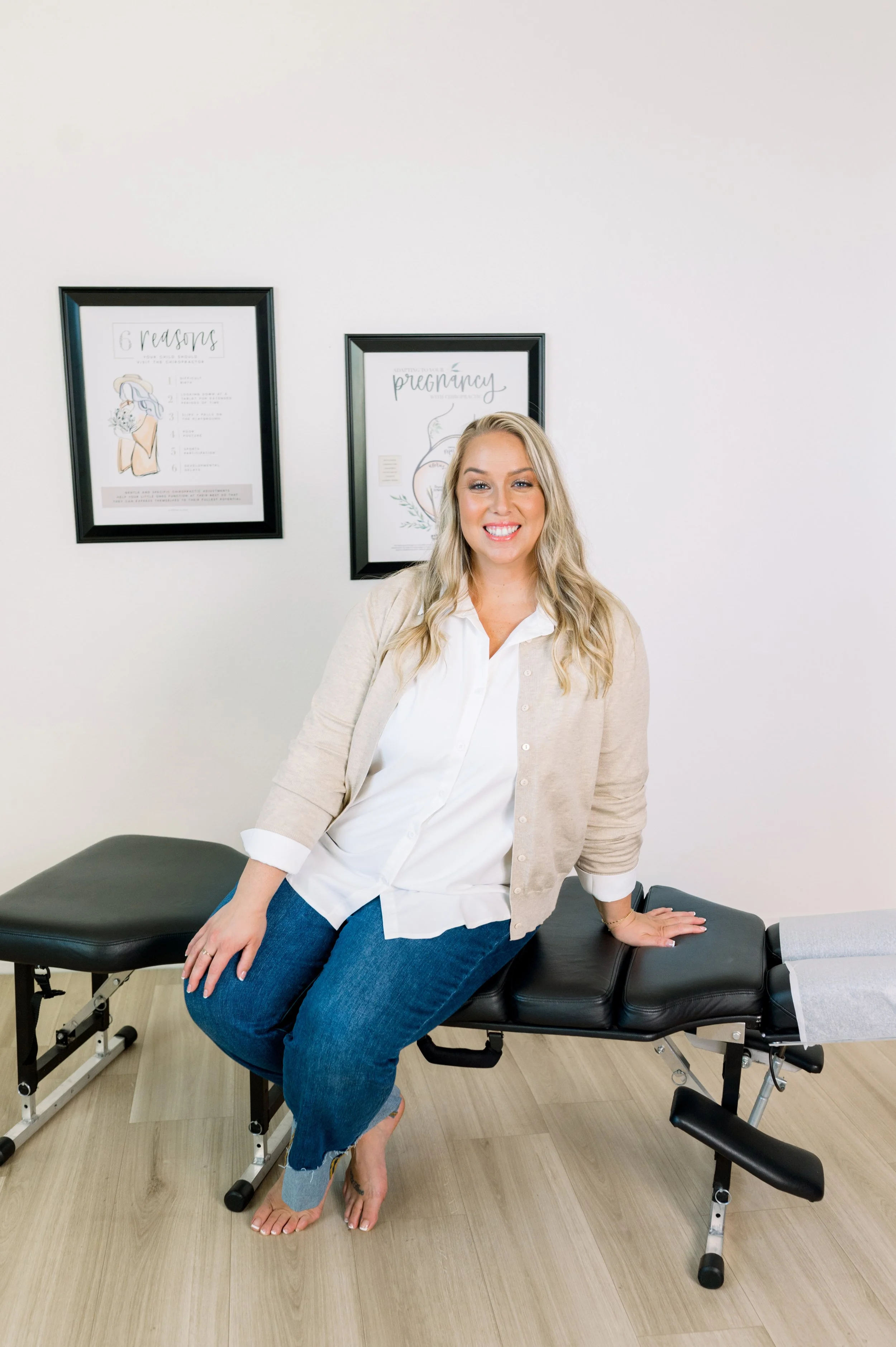 A smiling blonde woman sitting on a black chiropractic or massage table in a room with minimalist decor, including two framed posters on the wall behind her.