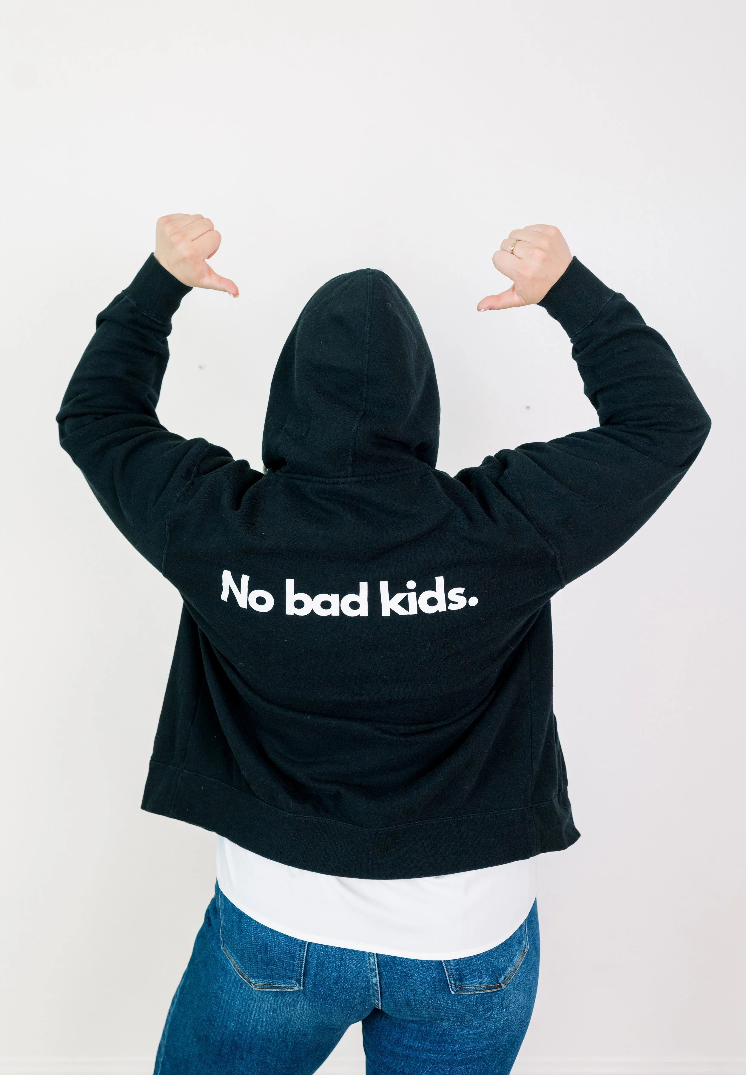 Person wearing a black hoodie with the wording 'No bad kids.' on the back, standing against a plain white wall, flexing arms.