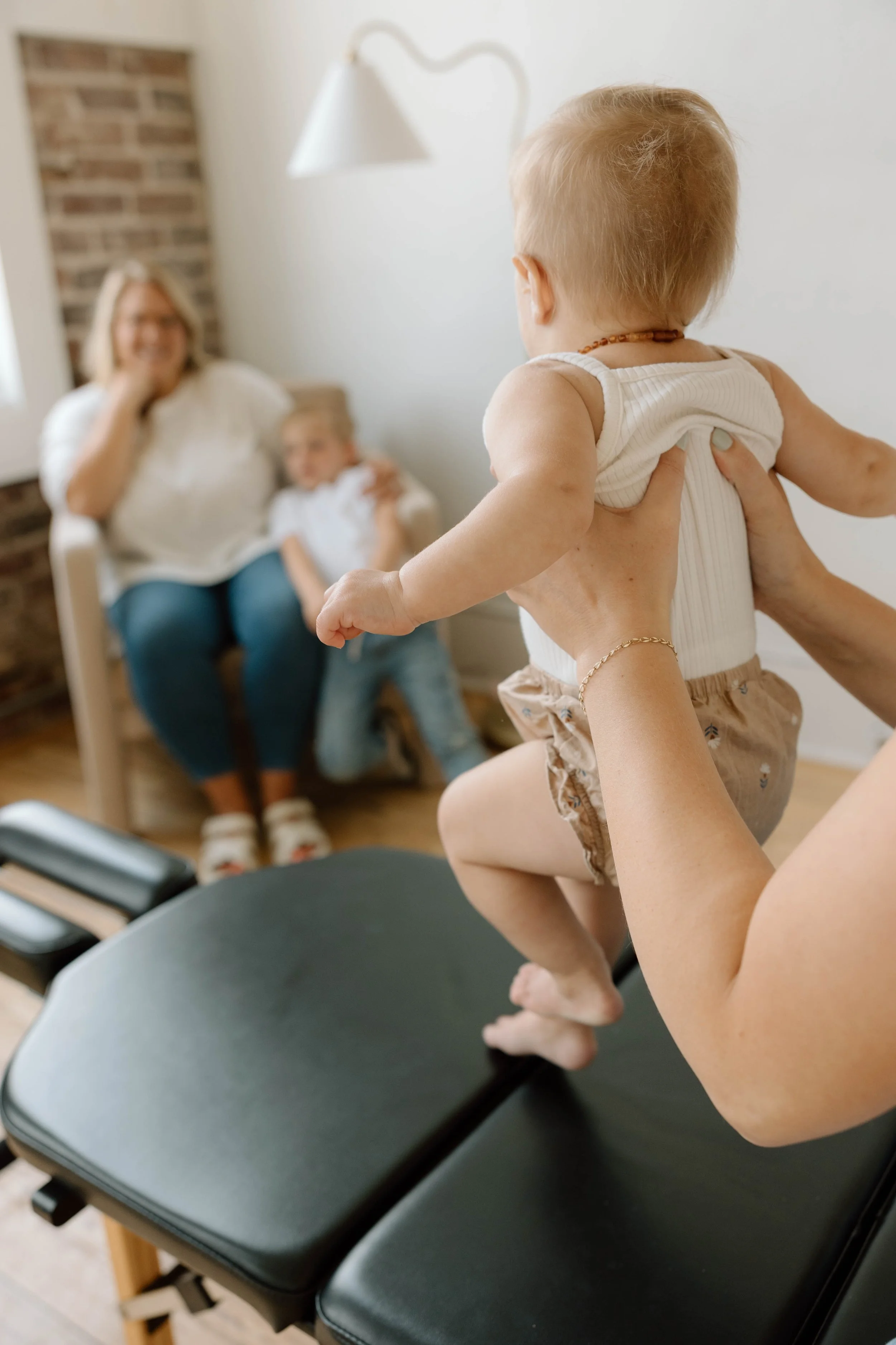 A baby being held up by a chiropractor, with a joyful woman and a young girl sitting on a chair in the background, inside a cozy room with a brick wall.