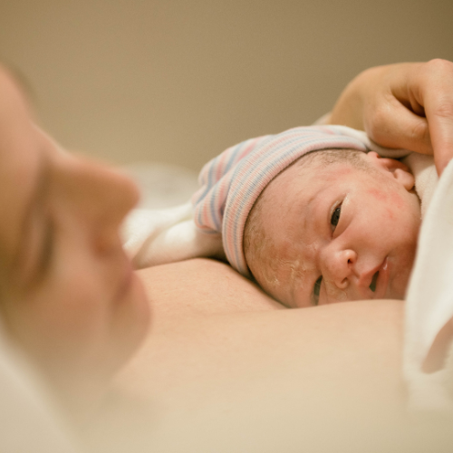 Newborn baby with a striped hat lying on their mom's chest in a hospital room.