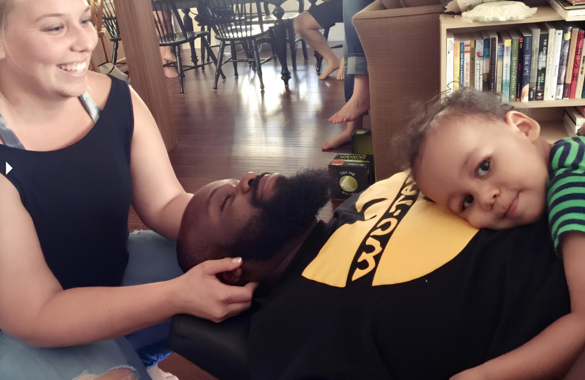 A smiling chiropractor checks a man's neck, while his baby with curly hair and a striped shirt lays on the man's belly, looking at the camera. 