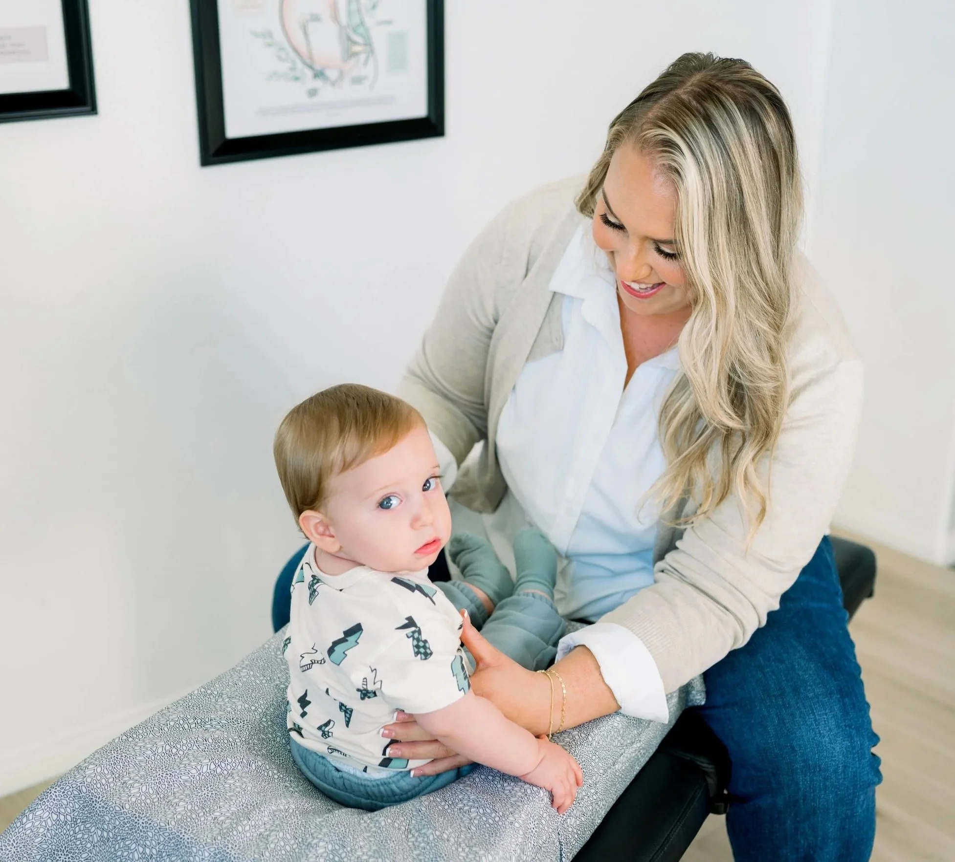 A woman smiling and looking down at a young boy, who is sitting on an examination table in a doctor's office. The woman appears to be a healthcare provider or a mother. The boy is wearing a white shirt with a pattern of animals and geometric shapes, looking at the camera with a neutral expression. The background shows framed medical illustrations on a white wall.
