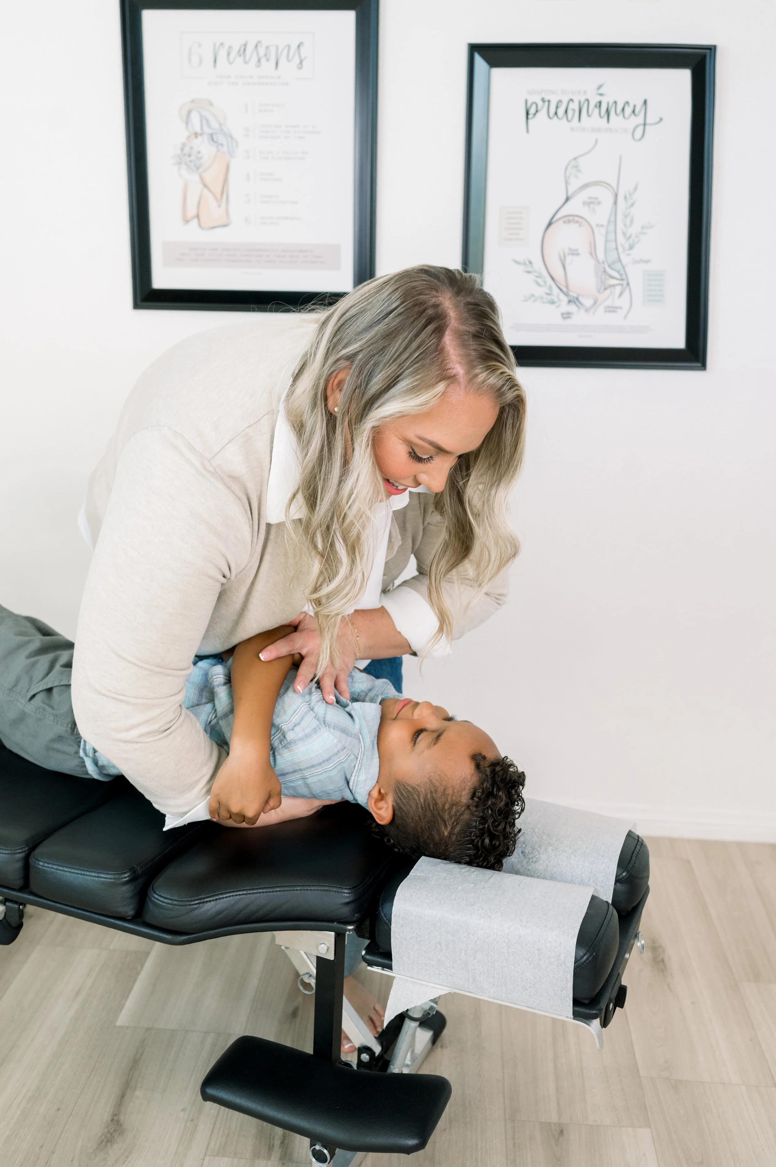 Female chiropractor adjusting a young boy lying on an adjustment table in a clinic with pregnancy and childbirth posters on the wall.