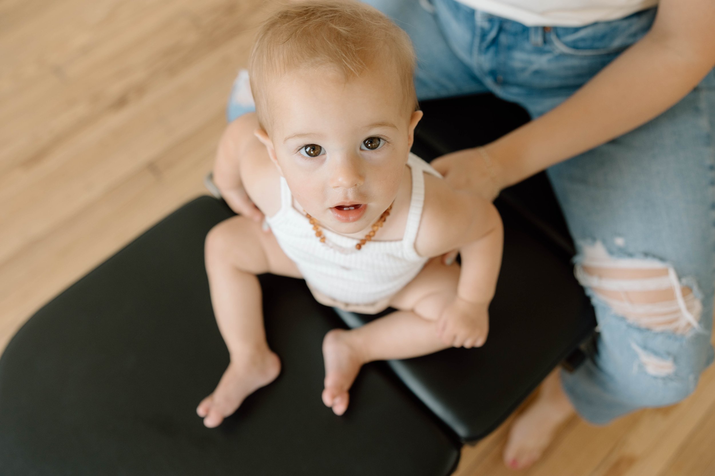 Young child sitting on a black chiropractic table, looking up at the camera, wearing a white sleeveless top and a teething necklace, with a chiropractor examining them.