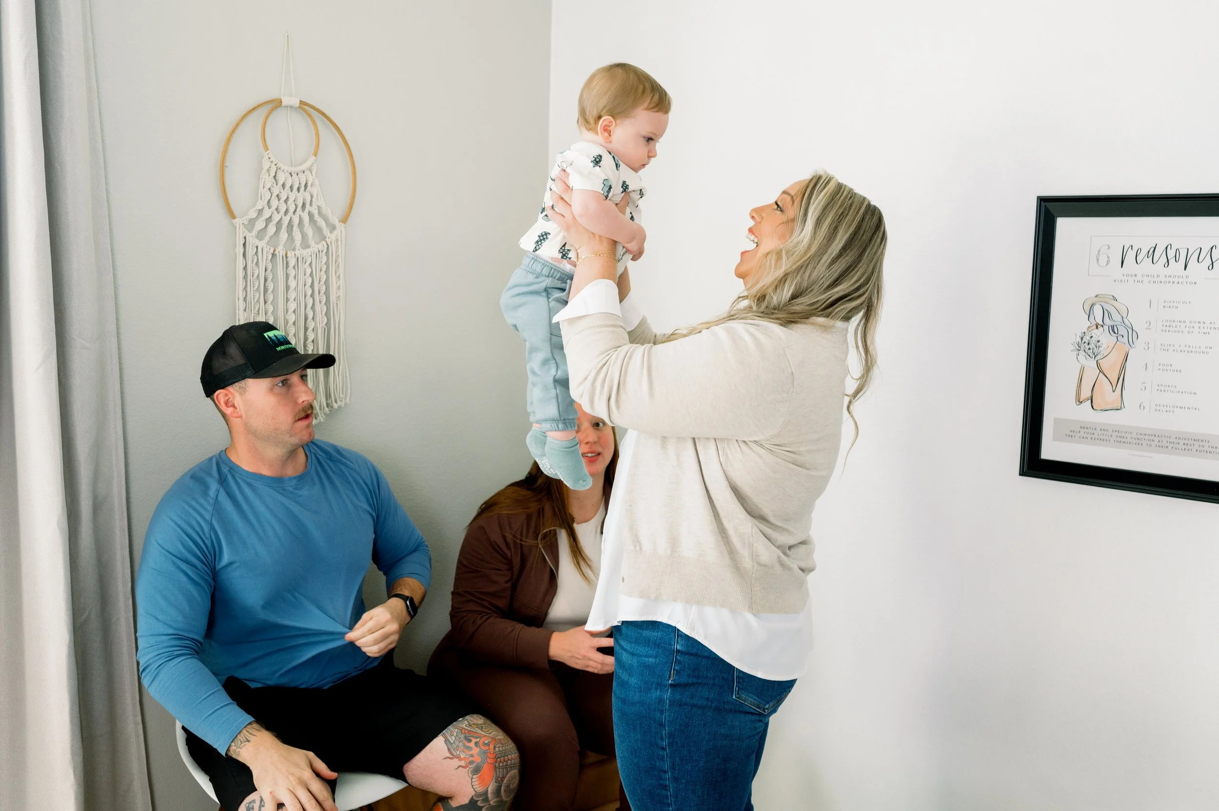 A woman lifts a young boy in the air, smiling, in a bright, modern room while two people watch. The woman has long blonde hair and is wearing a light-colored cardigan and jeans. A man with a tattoo on his leg, wearing a blue shirt and a cap, sits on a chair, and another woman with brown hair and a white shirt is seated beside him.