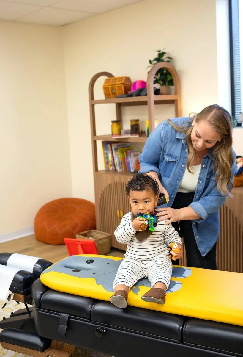Toddler sitting with toys on a pediatric chiropractic table while a chiropractor stands nearby at Taproot Chiropractic.