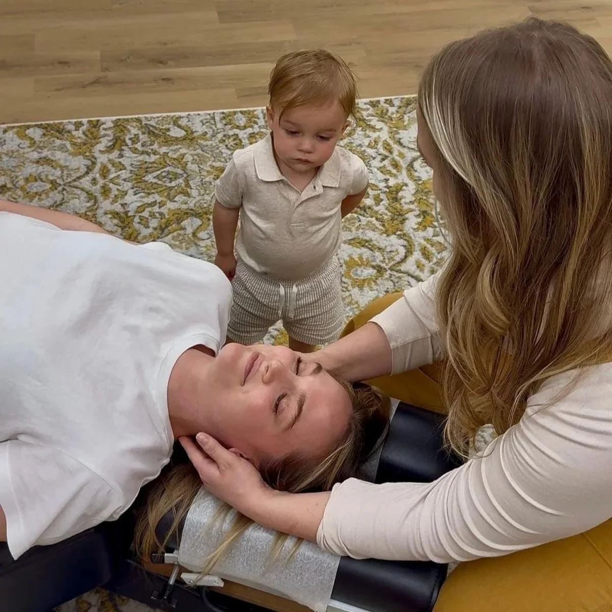 Dr. Jennifer Gordon gently adjusting a patient’s head and neck while a young child stands nearby.