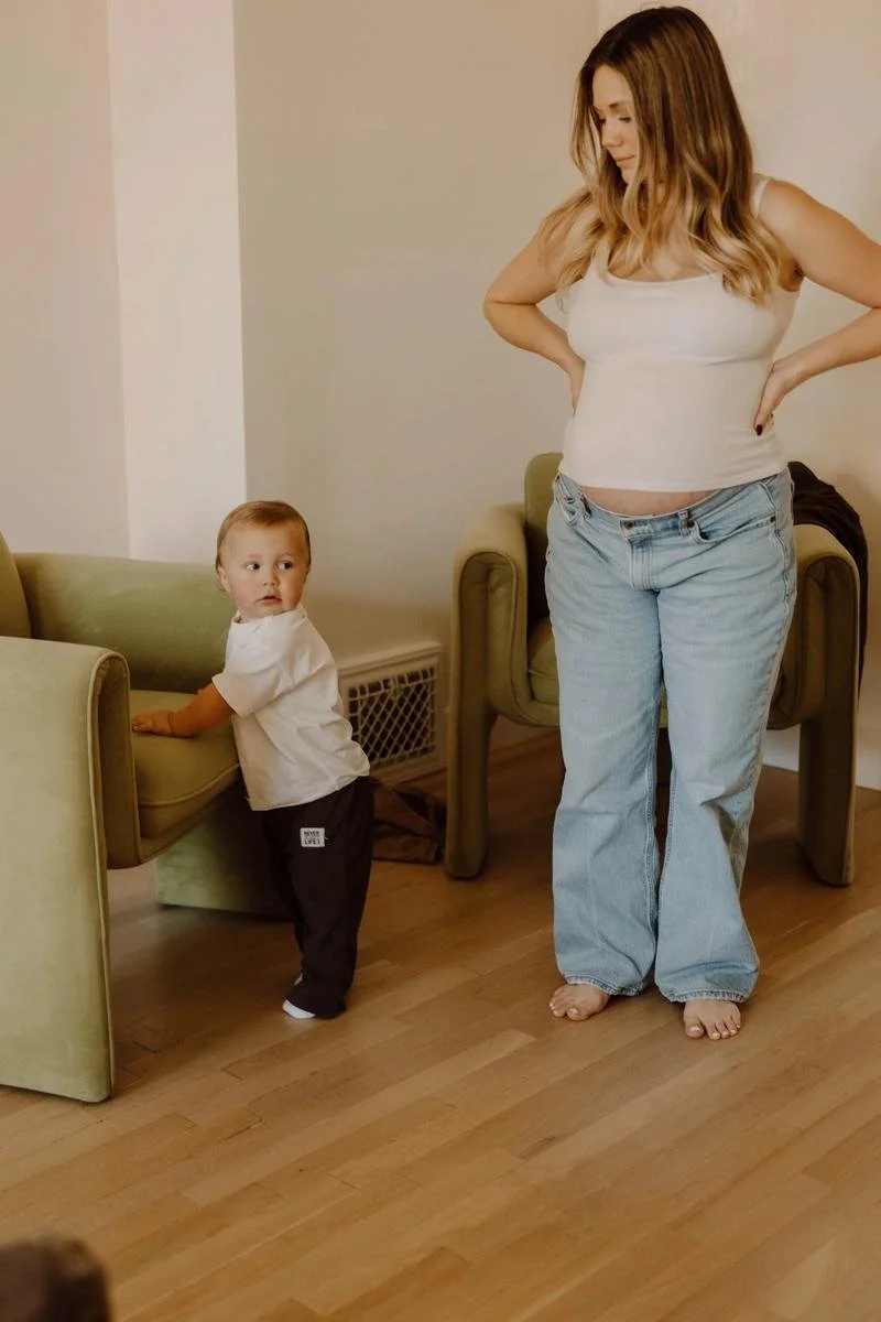 A pregnant patient standing with hands on her hips while a toddler plays nearby in the waiting room