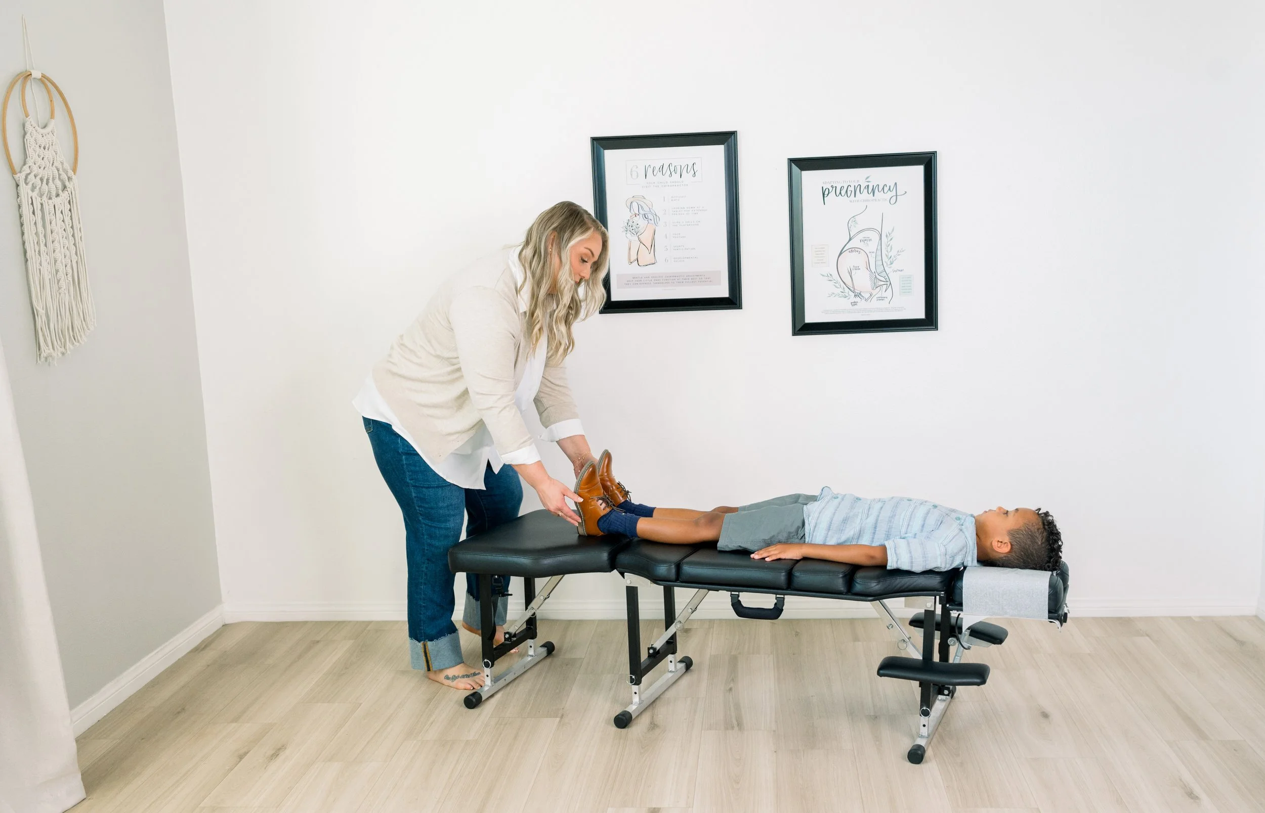 A chiropractor with long blonde hair aids a young boy with curly hair on a chiropractic table. The boy is lying down flat on his back while the woman holds his feet. The room is decorated with chiropractic posters hanging on the wall.