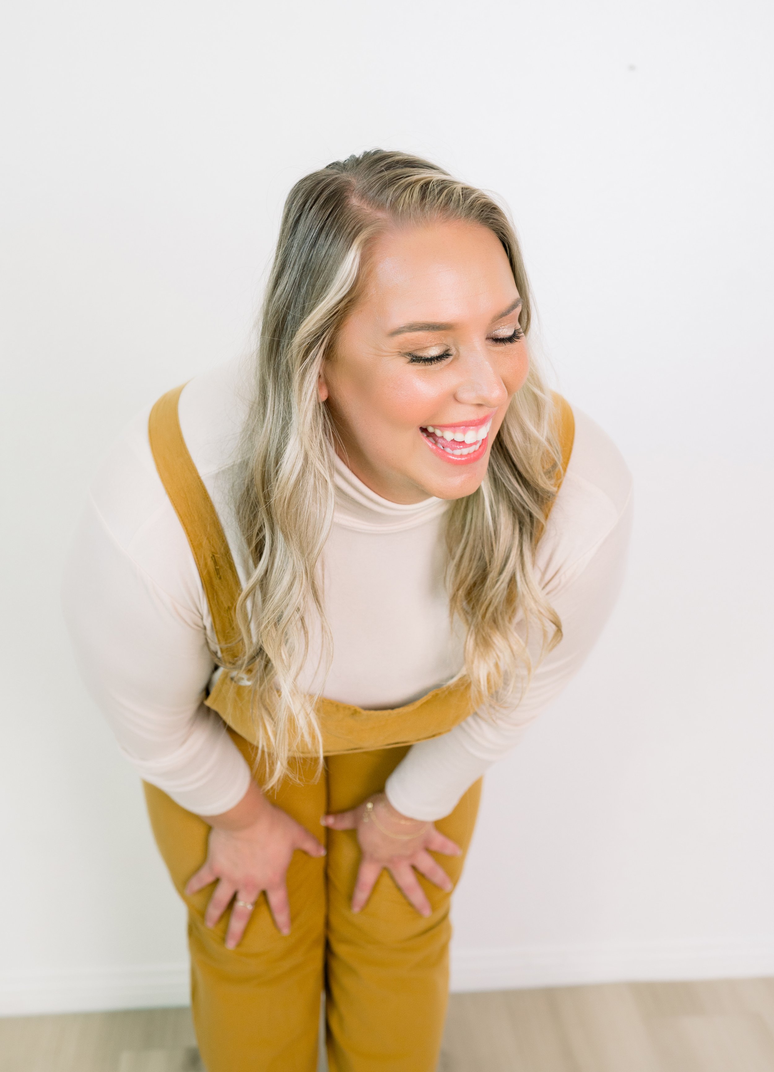 A woman with long blonde hair smiling and looking down, wearing a white top and mustard yellow overalls, standing against a white wall.