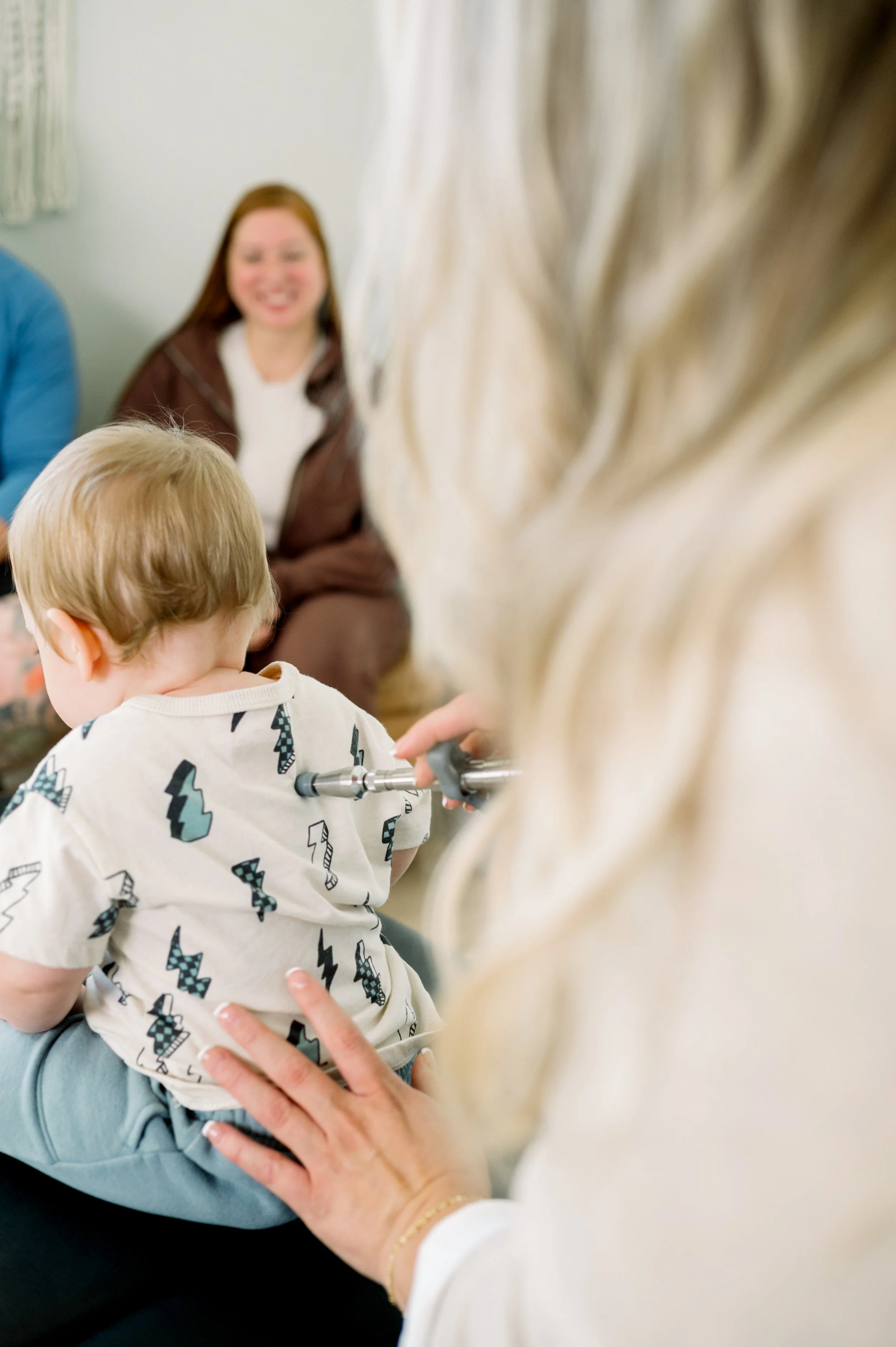 A young child with short light brown hair sitting on a chiropractic table while a chiropractor adjusts his lower spine with a low force adjusting tool in a chiropractic office, with a woman sitting in the background smiling.