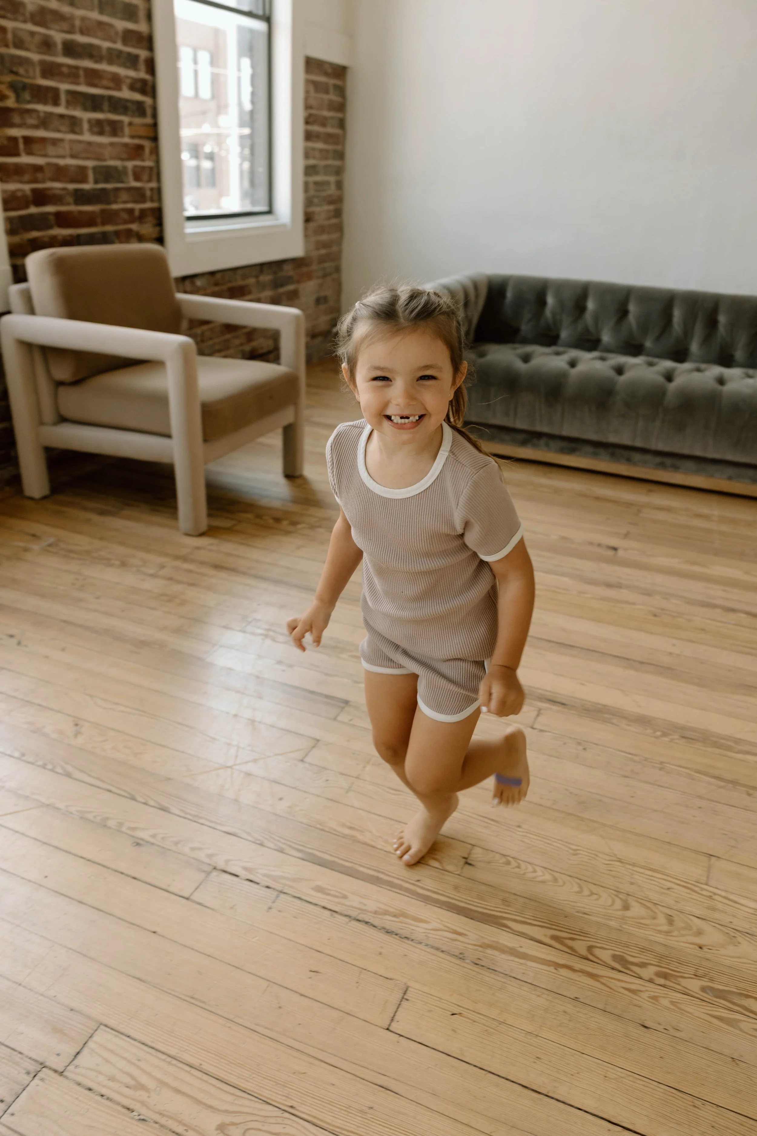 A young girl with a big smile, running and playing in a room with wooden floors, an armchair, and a sofa, with brick wall and window in the background.
