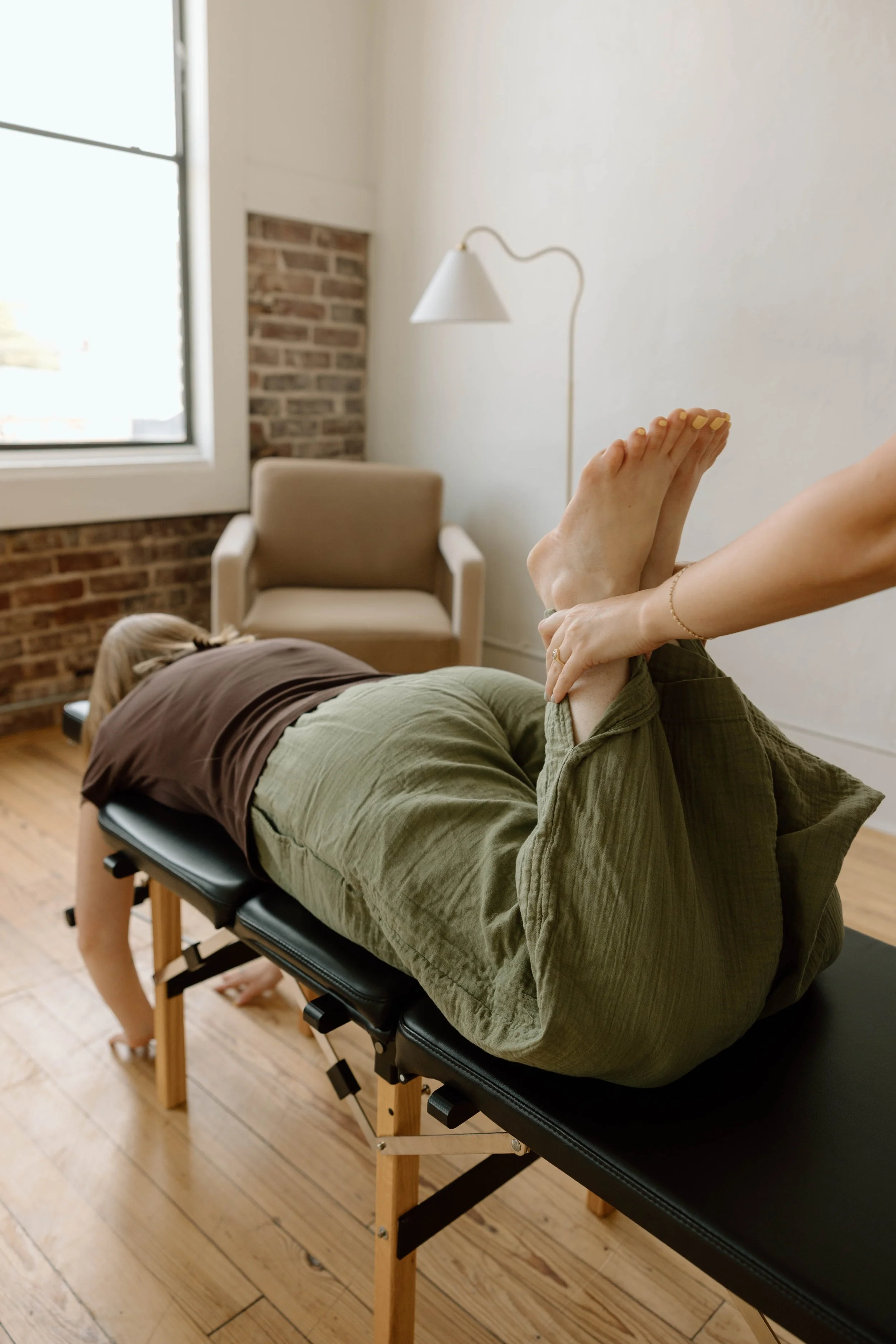 A person lying face down on a chiropractic table, receiving a leg check from a chiropractor in a chiropractic office with a window, a chair, and a wall lamp.