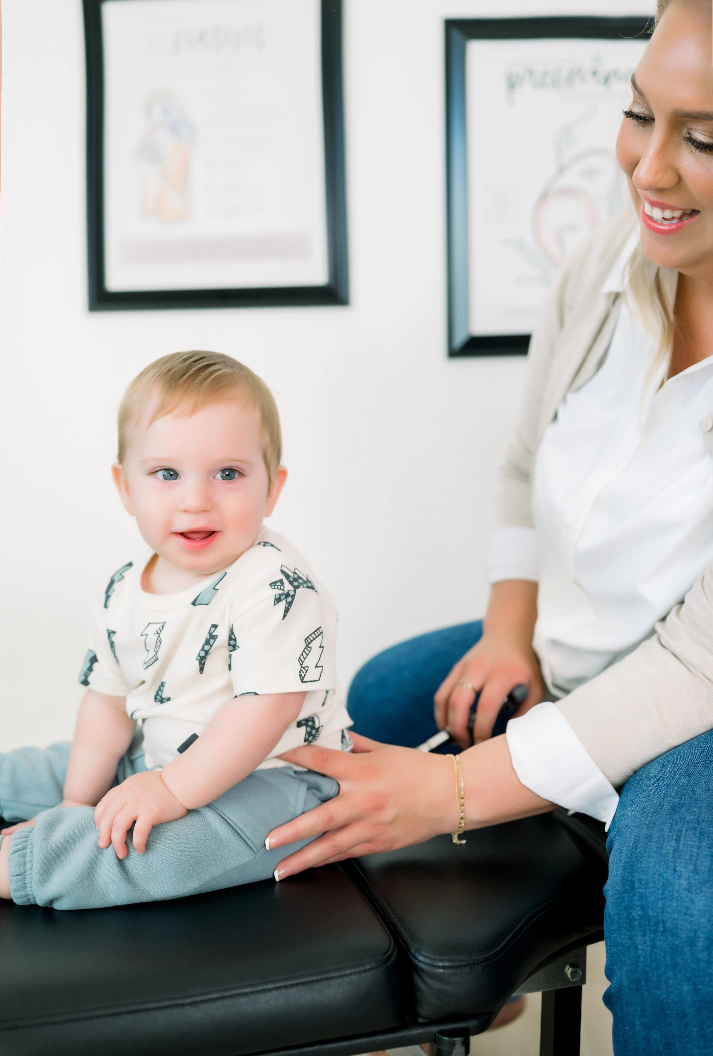 A young child sitting on an examination table at the doctor’s office with a smiling female healthcare provider beside him.
