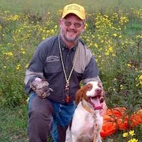 Photo of Tim Castillo in a blaze orange hunting cap with a Brittany hunting dog and a quail
