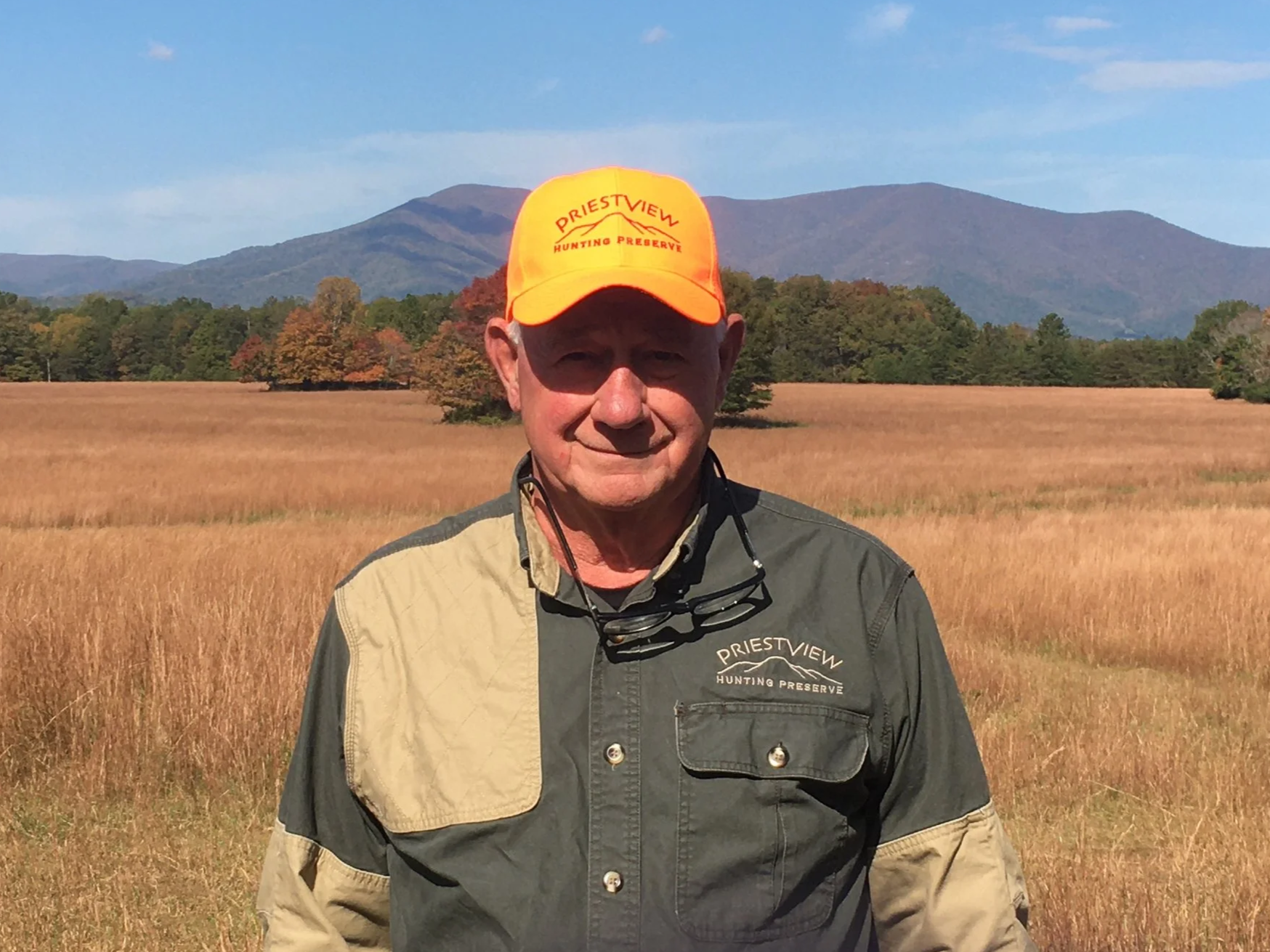 Photo of Bernie Davis in a blaze orange hat in front of a grass field with the Priest mountain in the background.