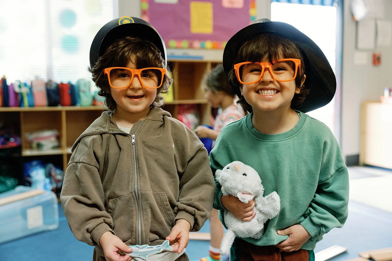 Two young boys dressed as magicians wearing black hats and large orange glasses, smiling in a classroom; one boy holding a stuffed animal, the other holding a magic wand.