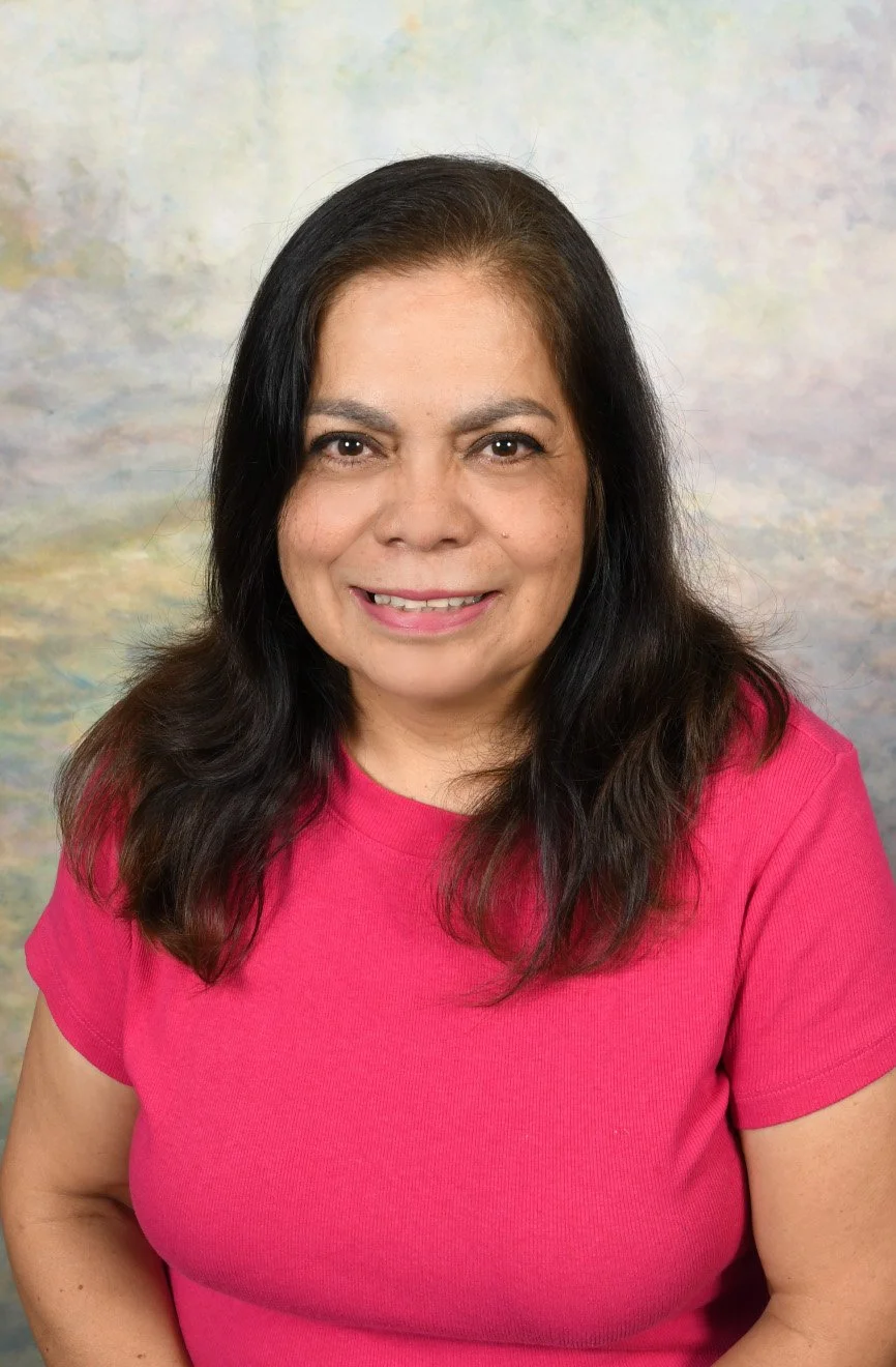 A woman with shoulder-length dark hair wearing a pink shirt, smiling, against a light-colored, textured background.