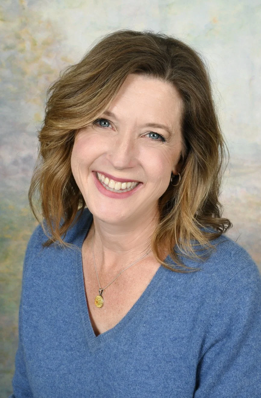 Portrait of a woman with shoulder-length wavy brown hair, blue eyes, wearing a blue V-neck sweater and a gold pendant necklace, smiling at the camera.