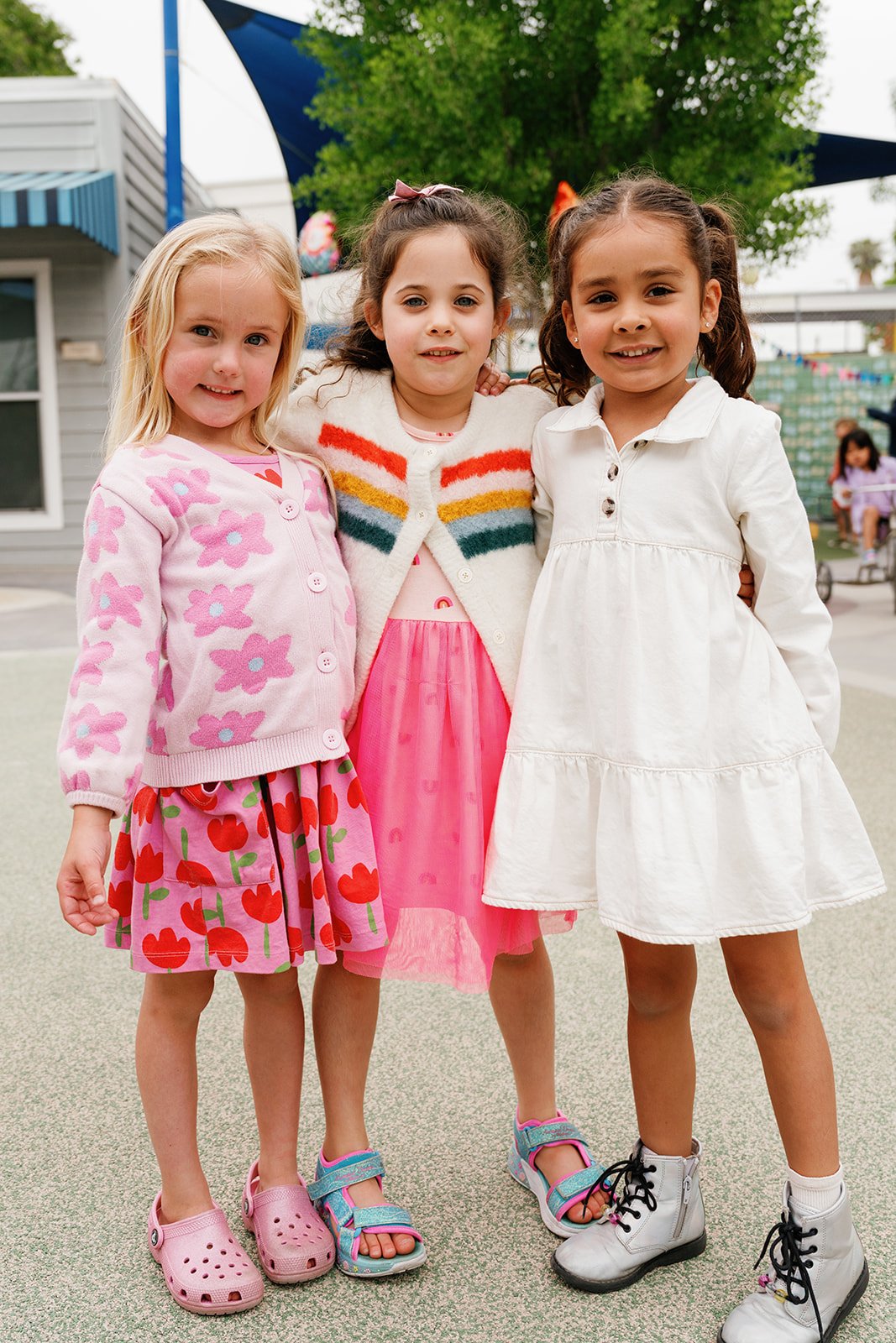 Three young girls standing together outdoors at a playground, smiling at the camera.