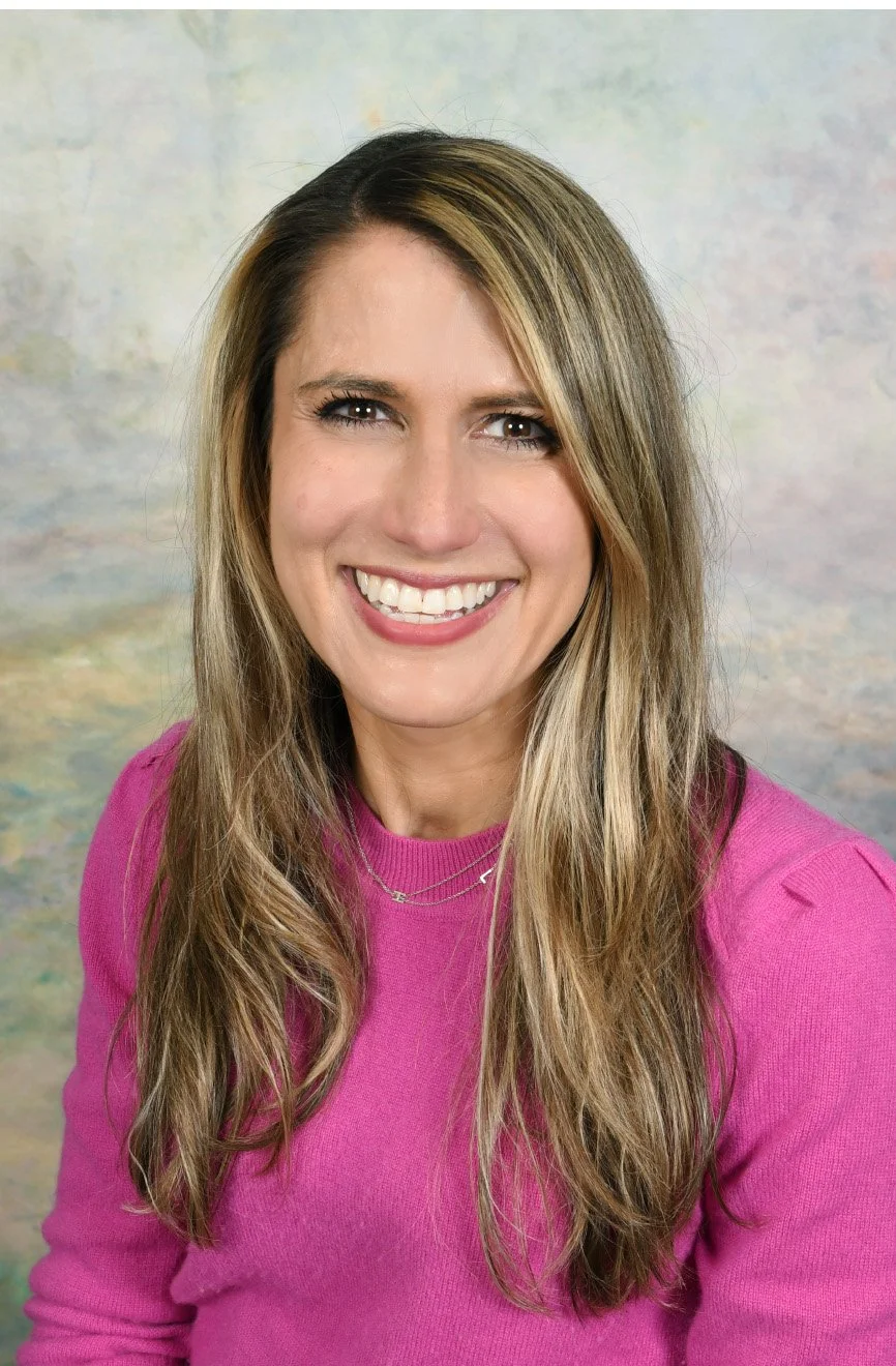 A smiling woman with long, wavy blonde hair wearing a pink top, posed against a neutral, textured background.