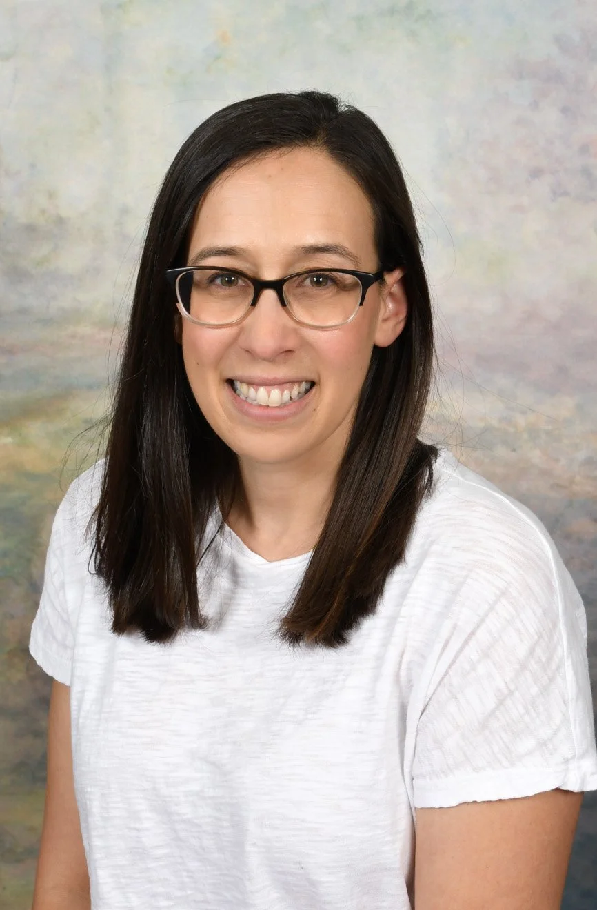 Portrait of a woman with long dark hair wearing glasses and a white t-shirt, smiling, against a colorful abstract background.