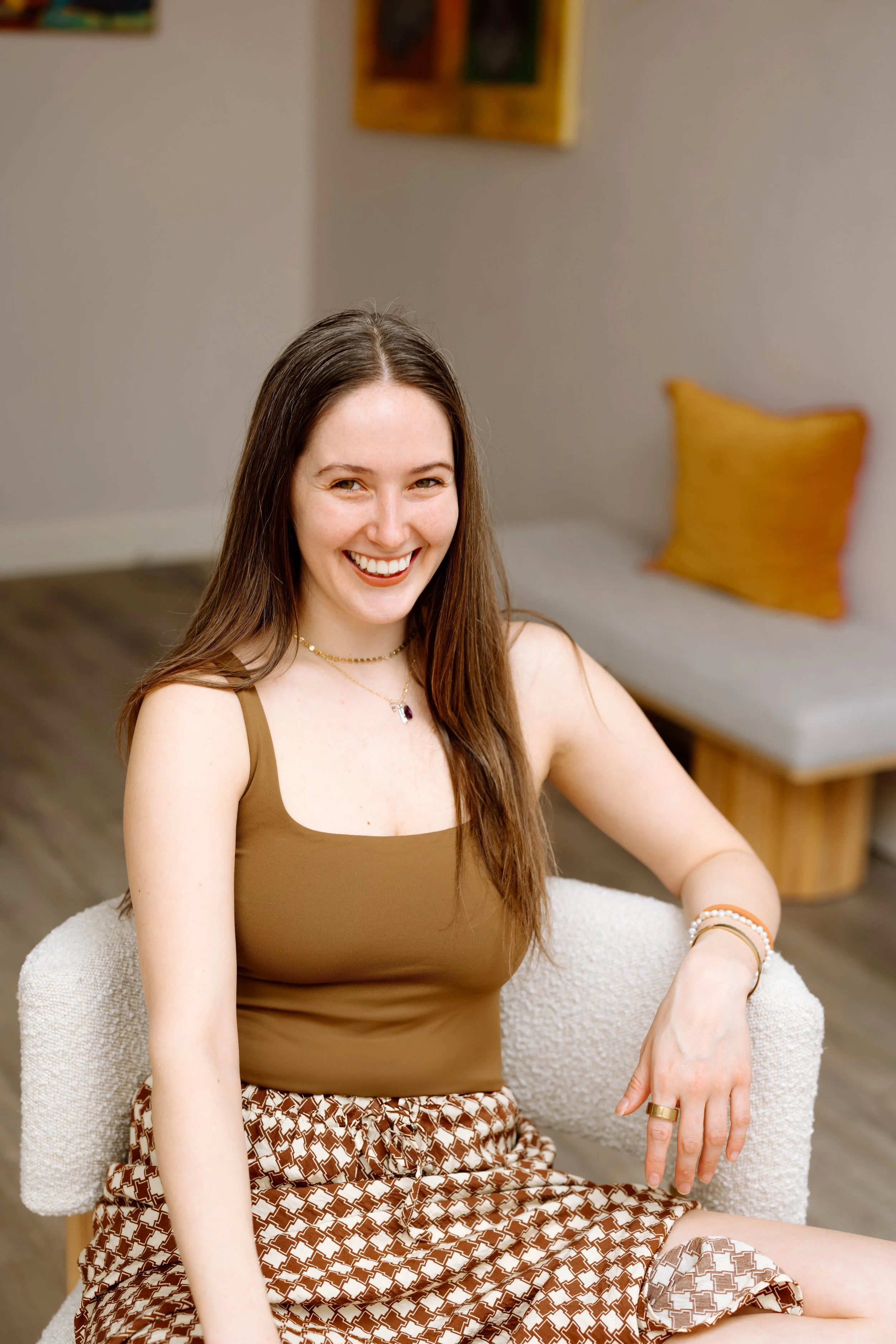 A young woman with long brown hair, wearing a brown tank top and patterned shorts, smiling and sitting on a cream-colored armchair in a cozy room with art on the walls.