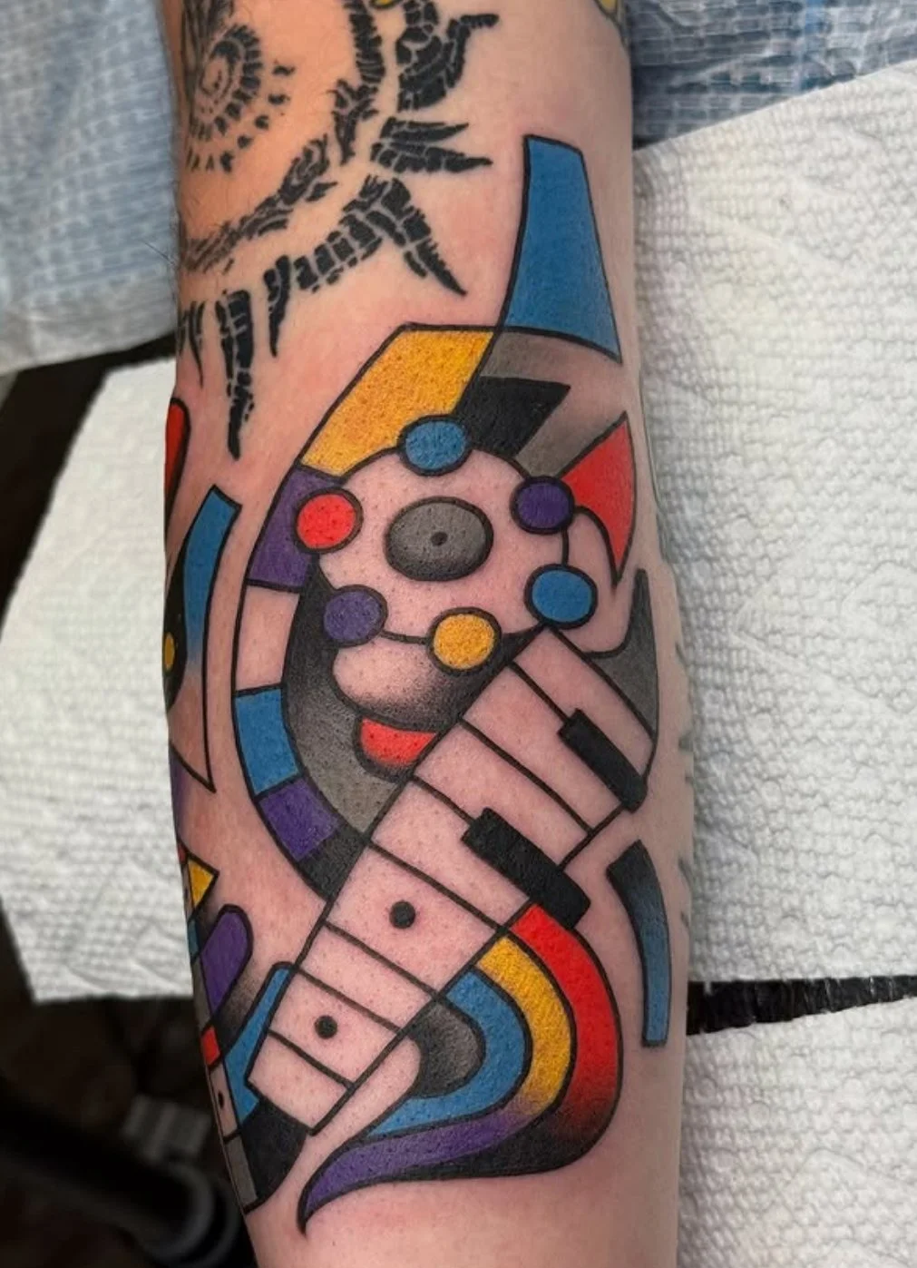 Colorful geometric tattoo featuring a stylized piano keyboard, musical notes, and abstract shapes with vibrant colors like red, blue, yellow, purple, and black.