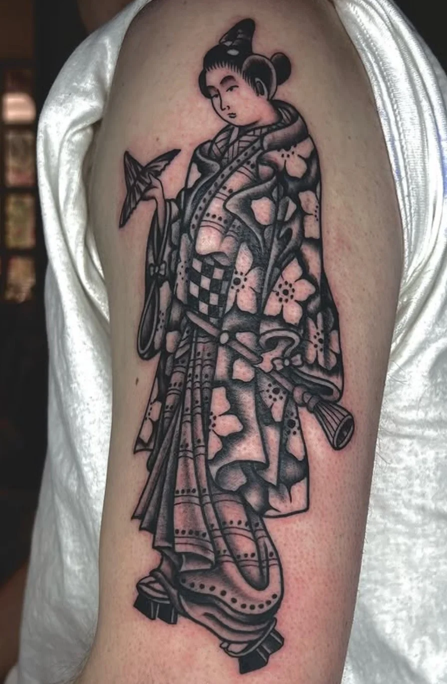 Tattoo of a traditionally styled Japanese geisha with dark hair, kimono with floral patterns, holding a fan, shown in black and gray ink.